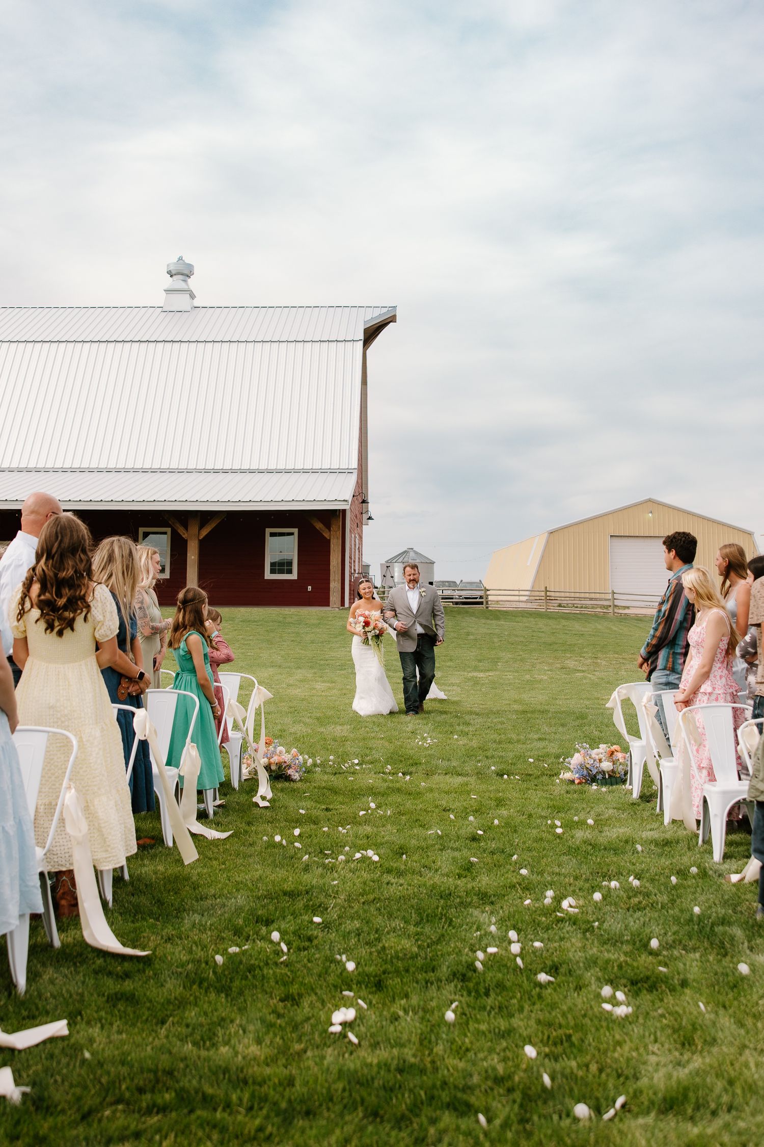 Bride walks down outdoor aisle with father at rustic farm wedding ceremony with white chairs.