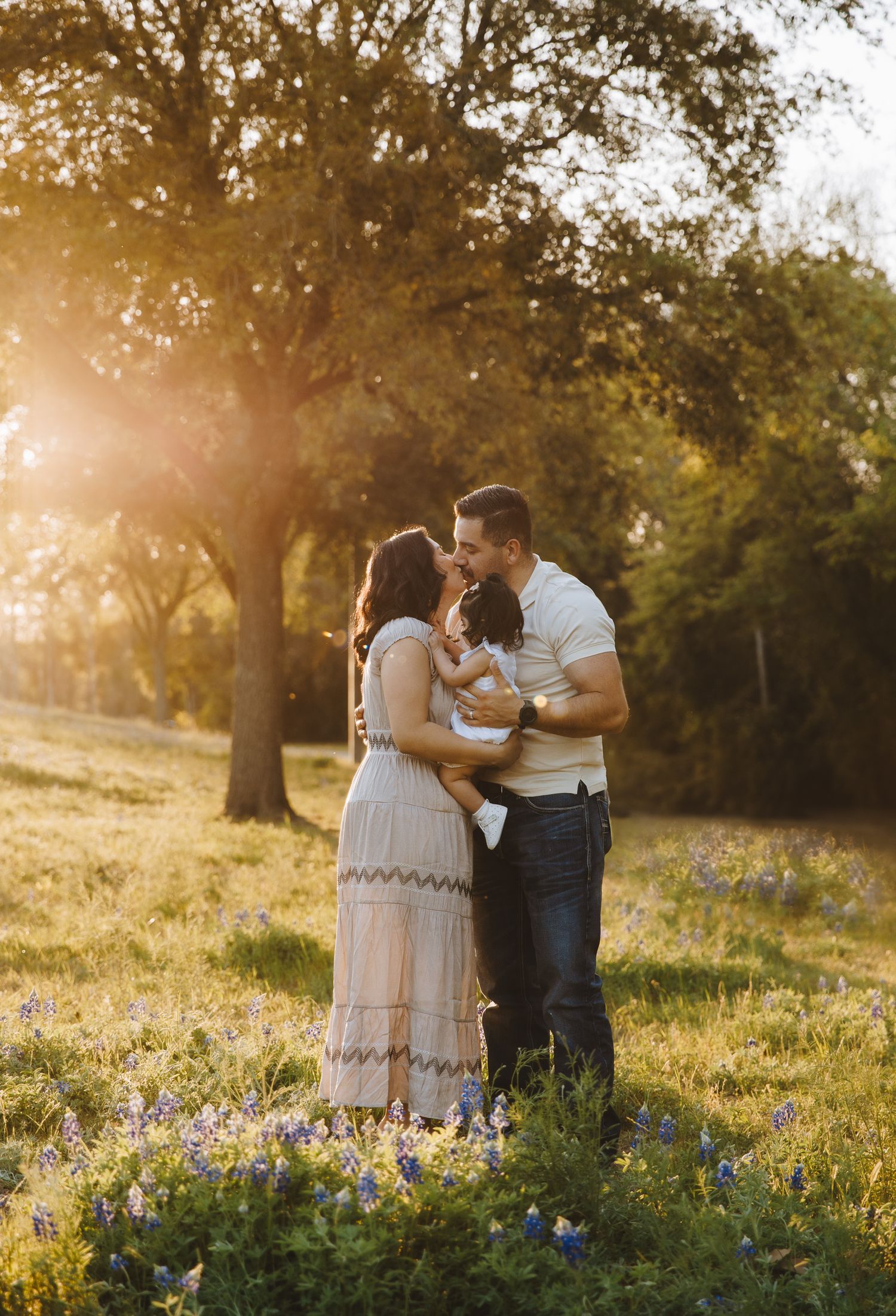 A couple shares a tender moment holding their baby in a sunlit wildflower meadow at golden hour.