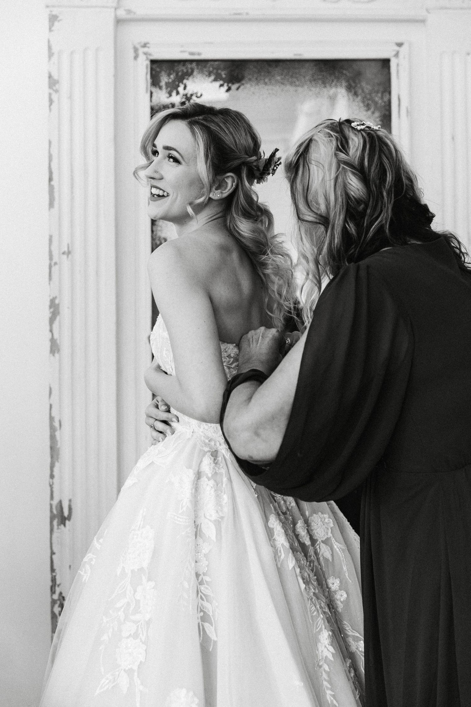 Black and white photo of laughing bride being helped into her wedding gown by a woman in a doorway.
