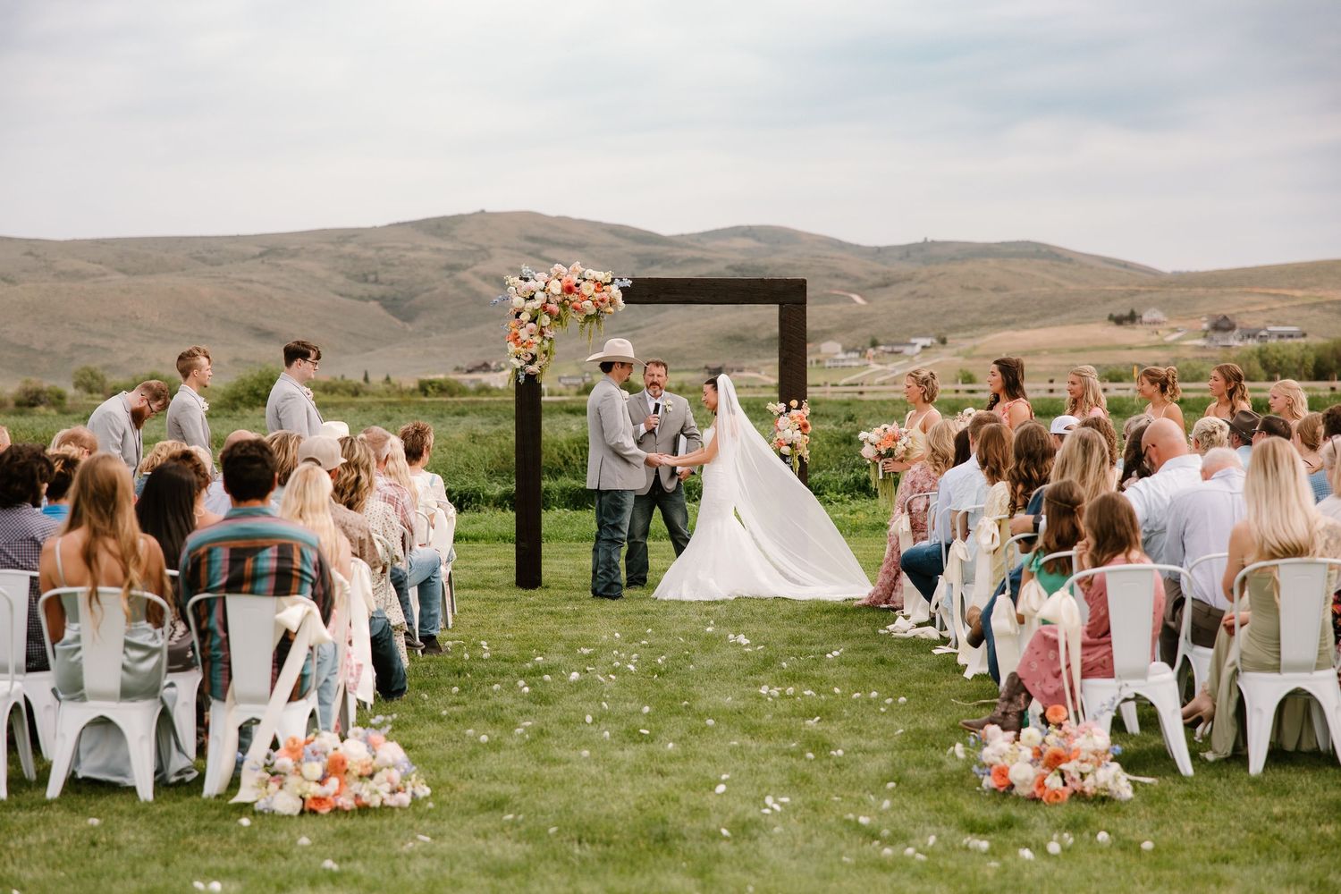 Outdoor wedding ceremony on green lawn with floral arch, guests seated in white chairs, rolling hills backdrop.