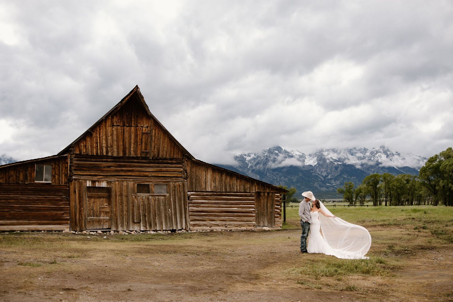 Bride and groom kiss near a historic wooden barn with mountain backdrop.