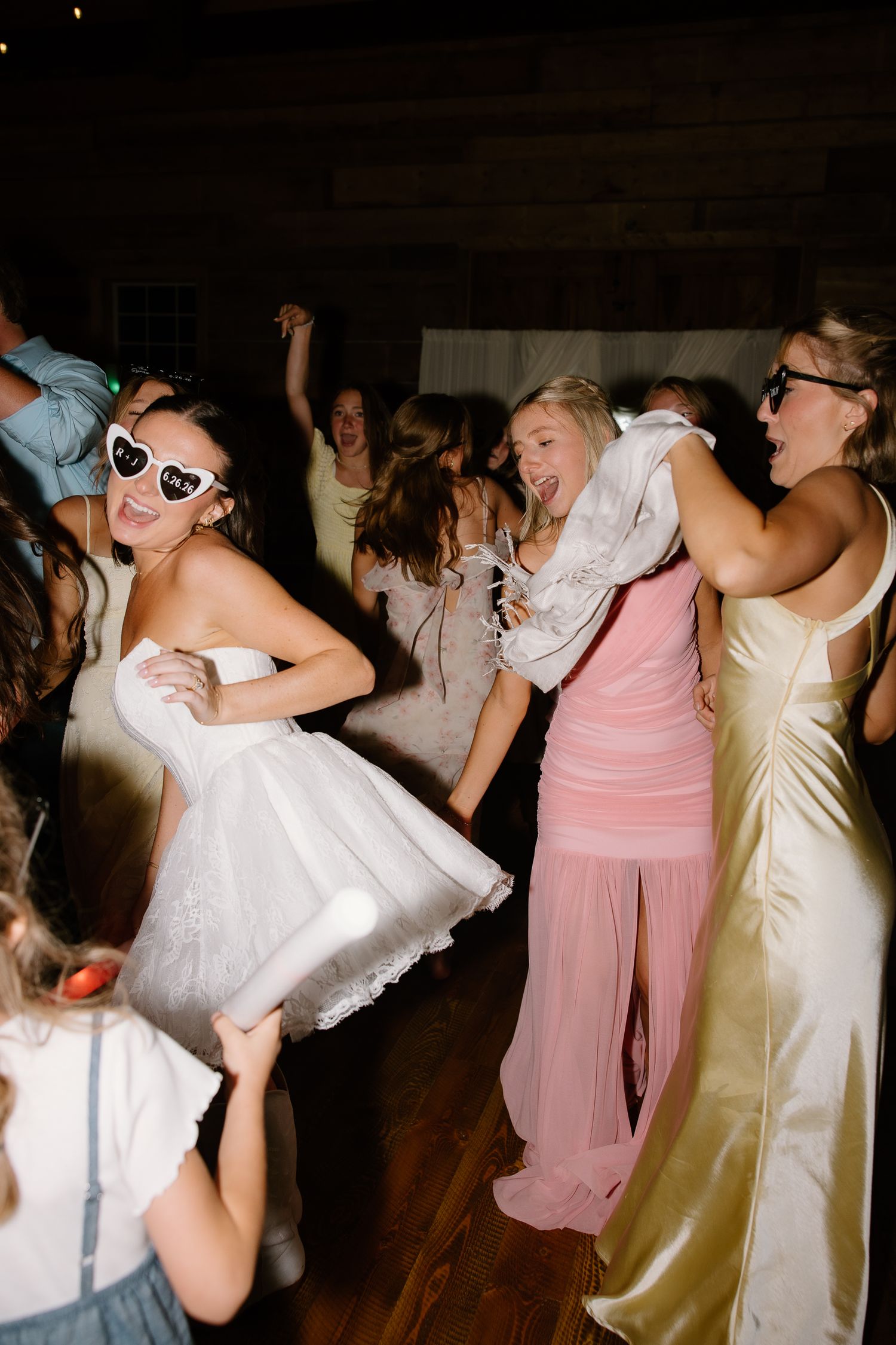 Bride in short white dress and heart sunglasses dancing energetically with guests at wedding reception.