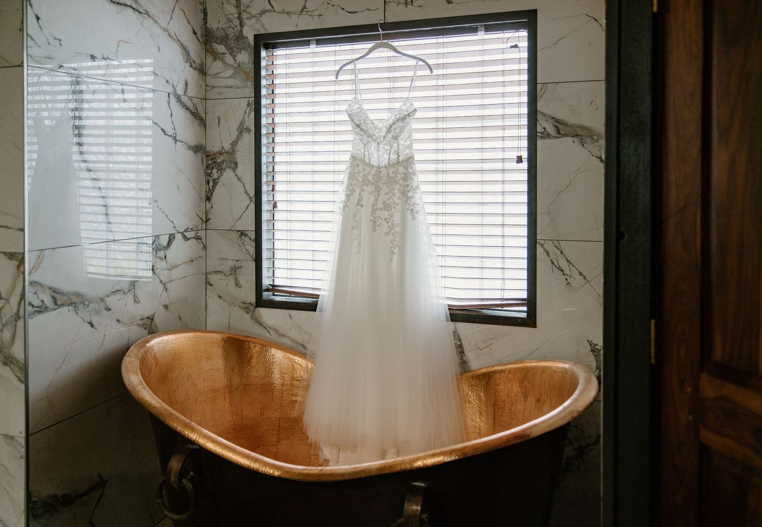 Elegant lace wedding dress hanging on a hanger over a copper clawfoot bathtub in a marble bathroom.