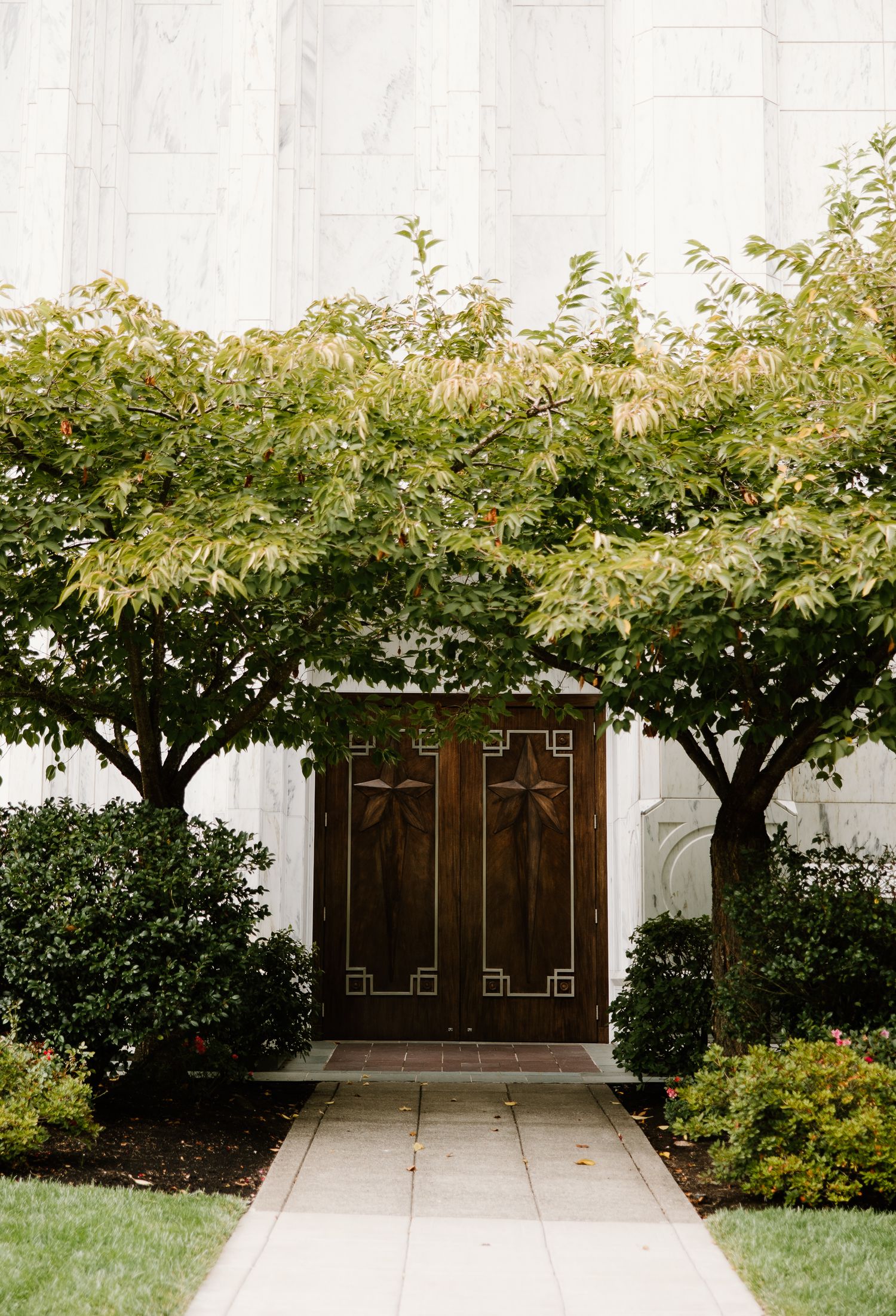 Ornate wooden double doors framed by lush green trees and manicured shrubs at a white stone building entrance.