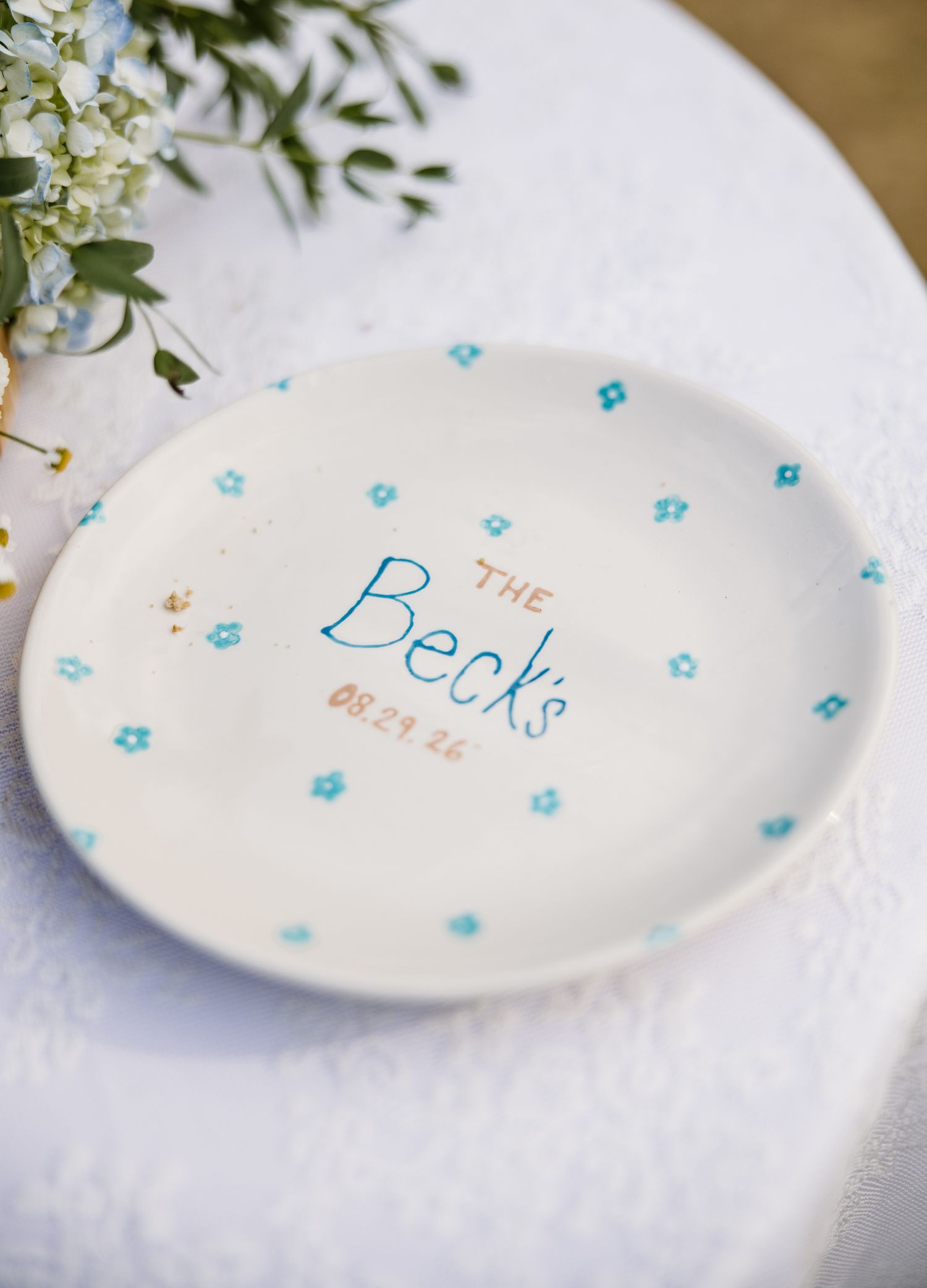 Handpainted ceramic plate reading The Becks 08.29.22 with blue floral details on a white tablecloth.