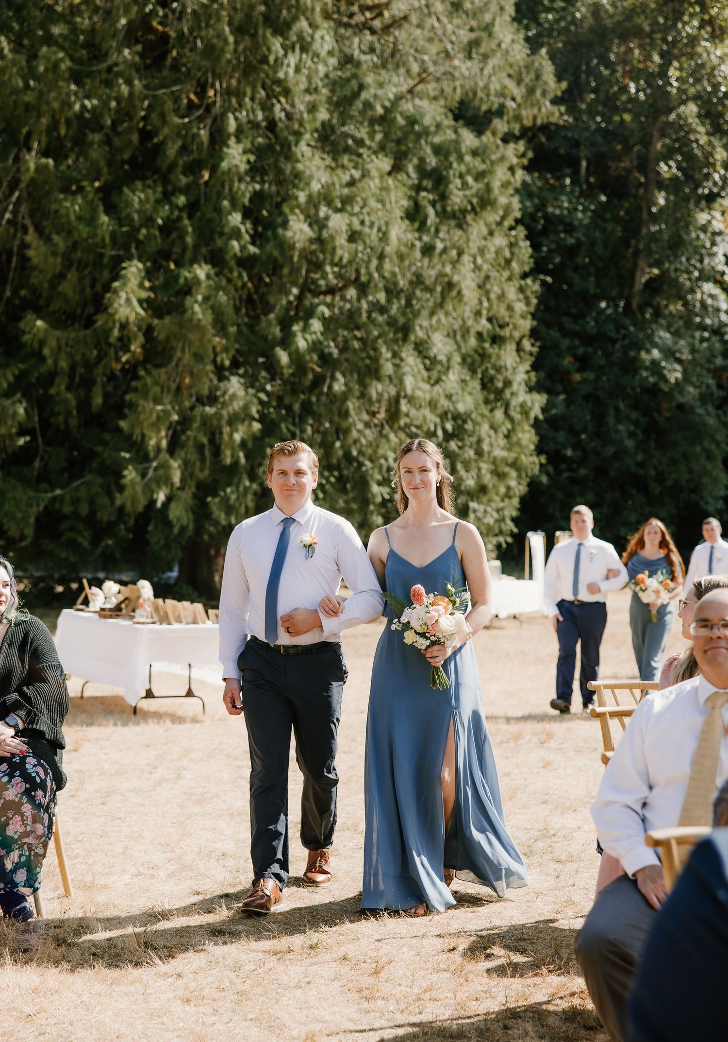 Bridesmaid in blue dress walks down outdoor wedding aisle with groomsman in navy suit.