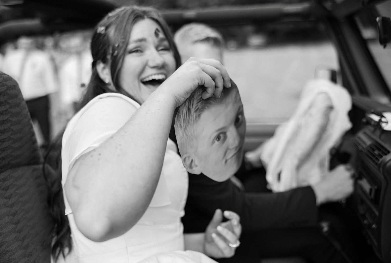 A laughing bride in white holds a young boy inside a vintage vehicle in a black and white photo.