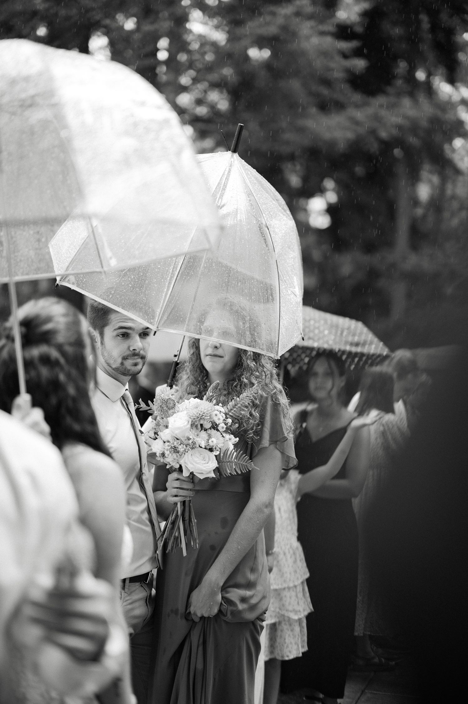 Black and white photo of a bride holding a bouquet under a clear umbrella in the rain at an outdoor wedding ceremony.