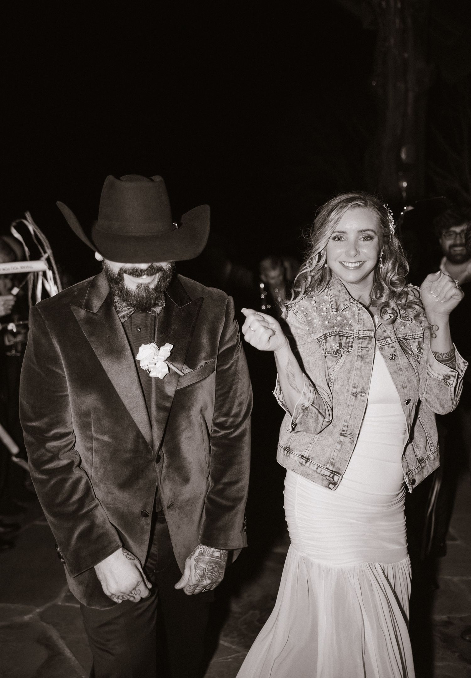 Black and white photo of newlyweds exiting reception, bride in denim jacket, groom in cowboy hat.