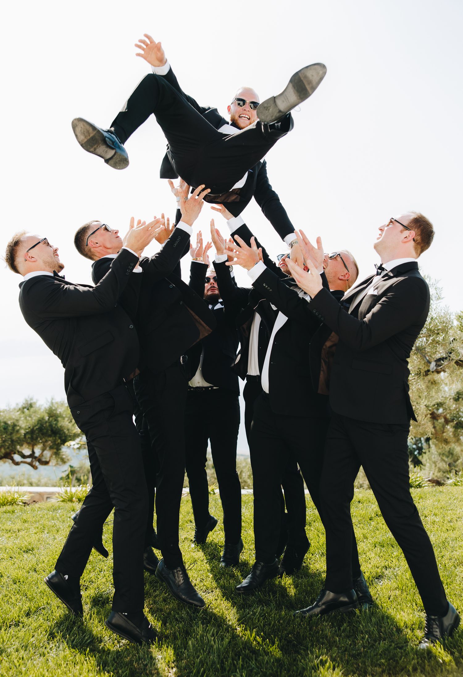 Groomsmen in black tuxedos toss the groom into the air outdoors on a sunny day in a celebratory moment.