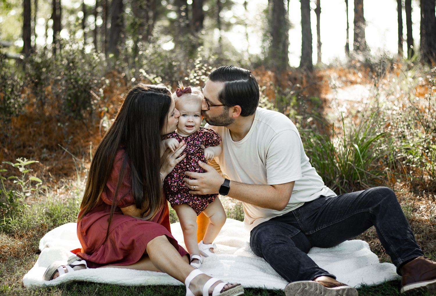 Young couple sitting on a blanket in a sunlit forest, sharing a kiss over their smiling baby girl.