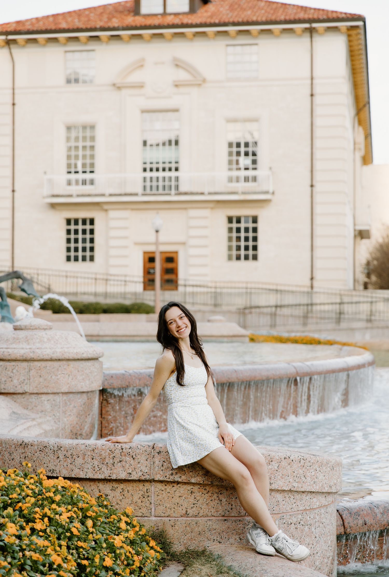 Smiling young woman in a white dress sitting by a fountain outside a classic university building.