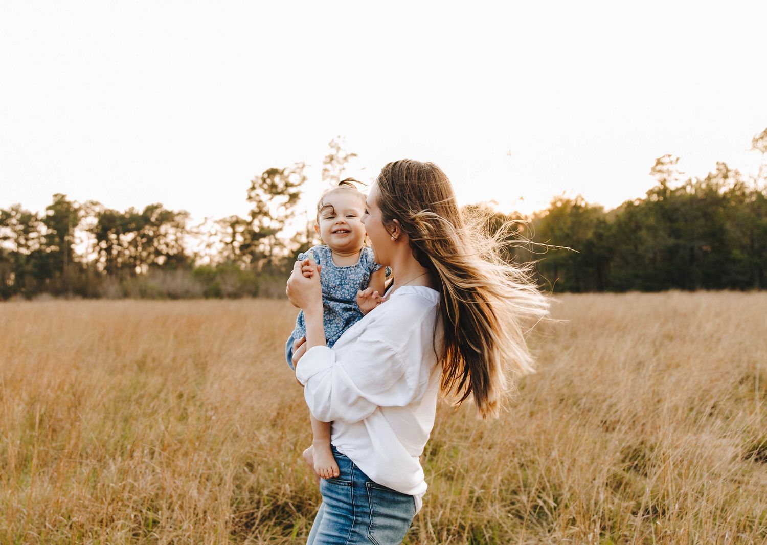 Joyful mother holds laughing baby girl in golden sunlit field during outdoor family photo session.