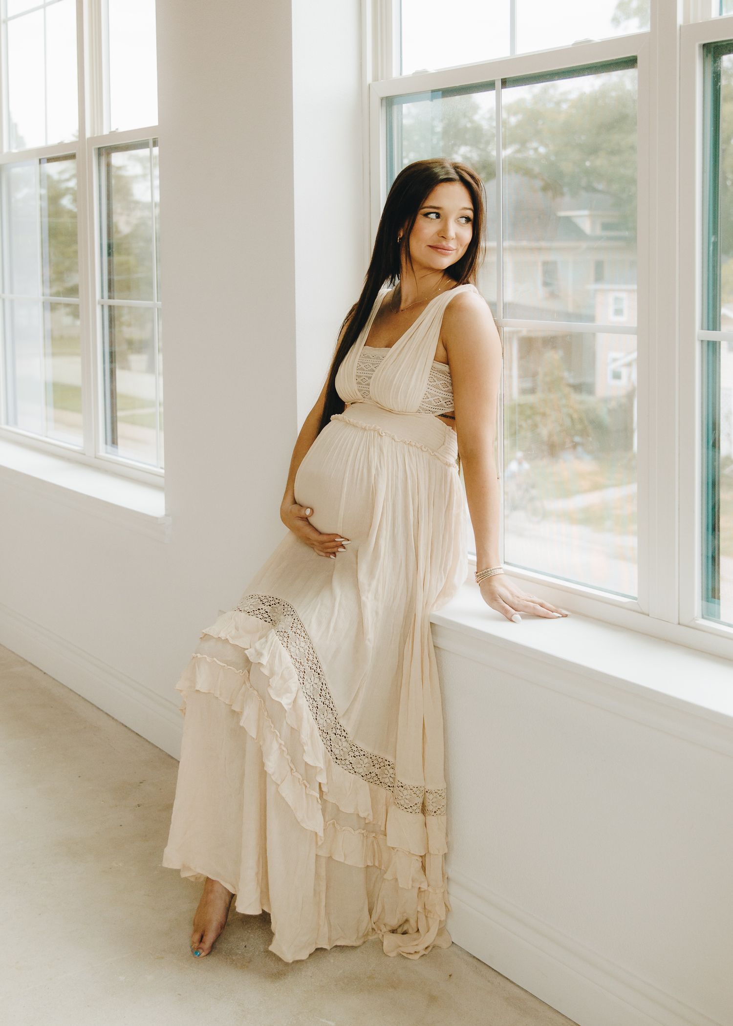 Pregnant woman in elegant cream boho maxi dress stands by window holding baby bump during maternity session.