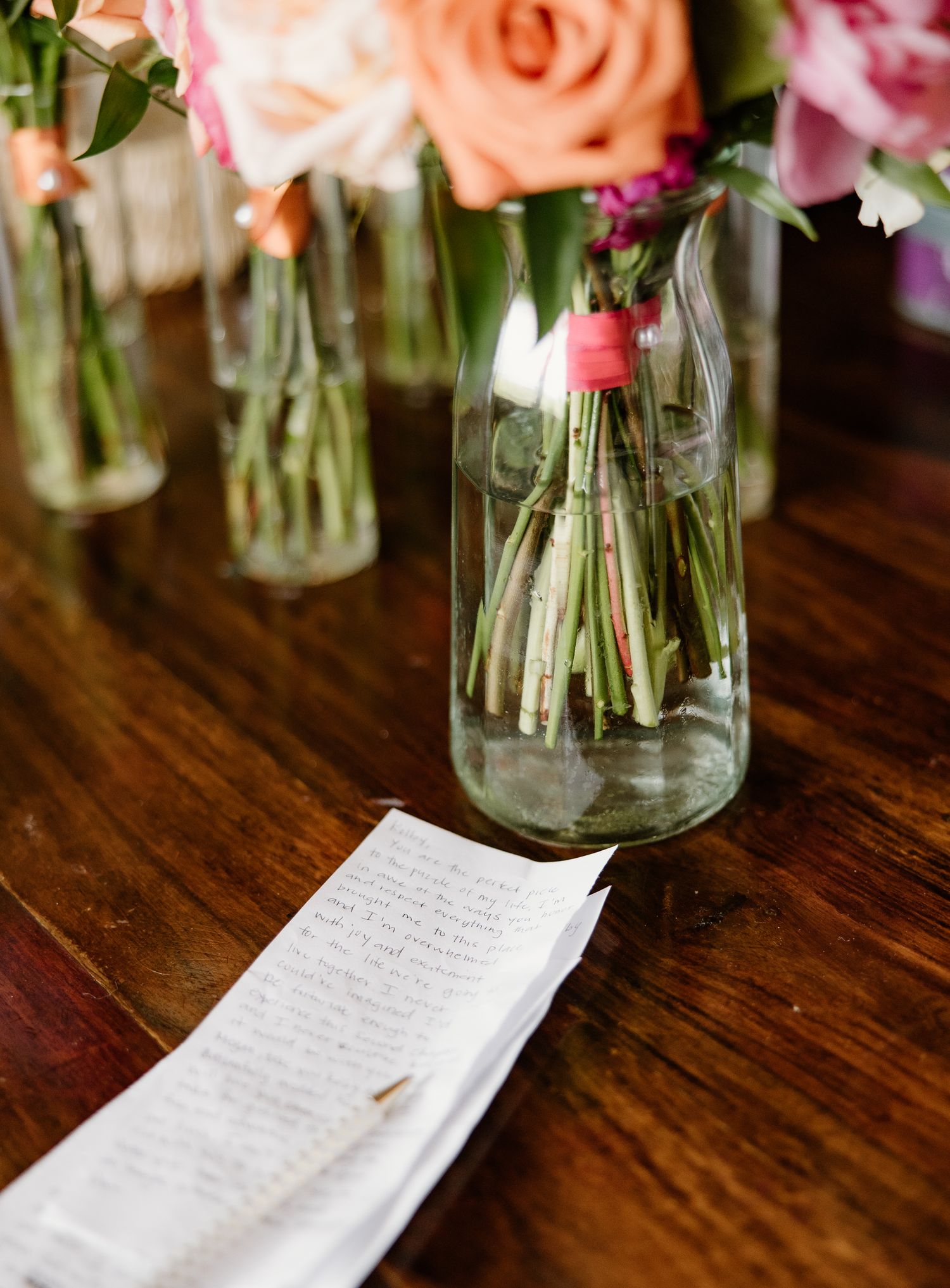 Glass bottle vases with fresh flowers and greenery on a wooden table beside a handwritten note at a wedding reception.
