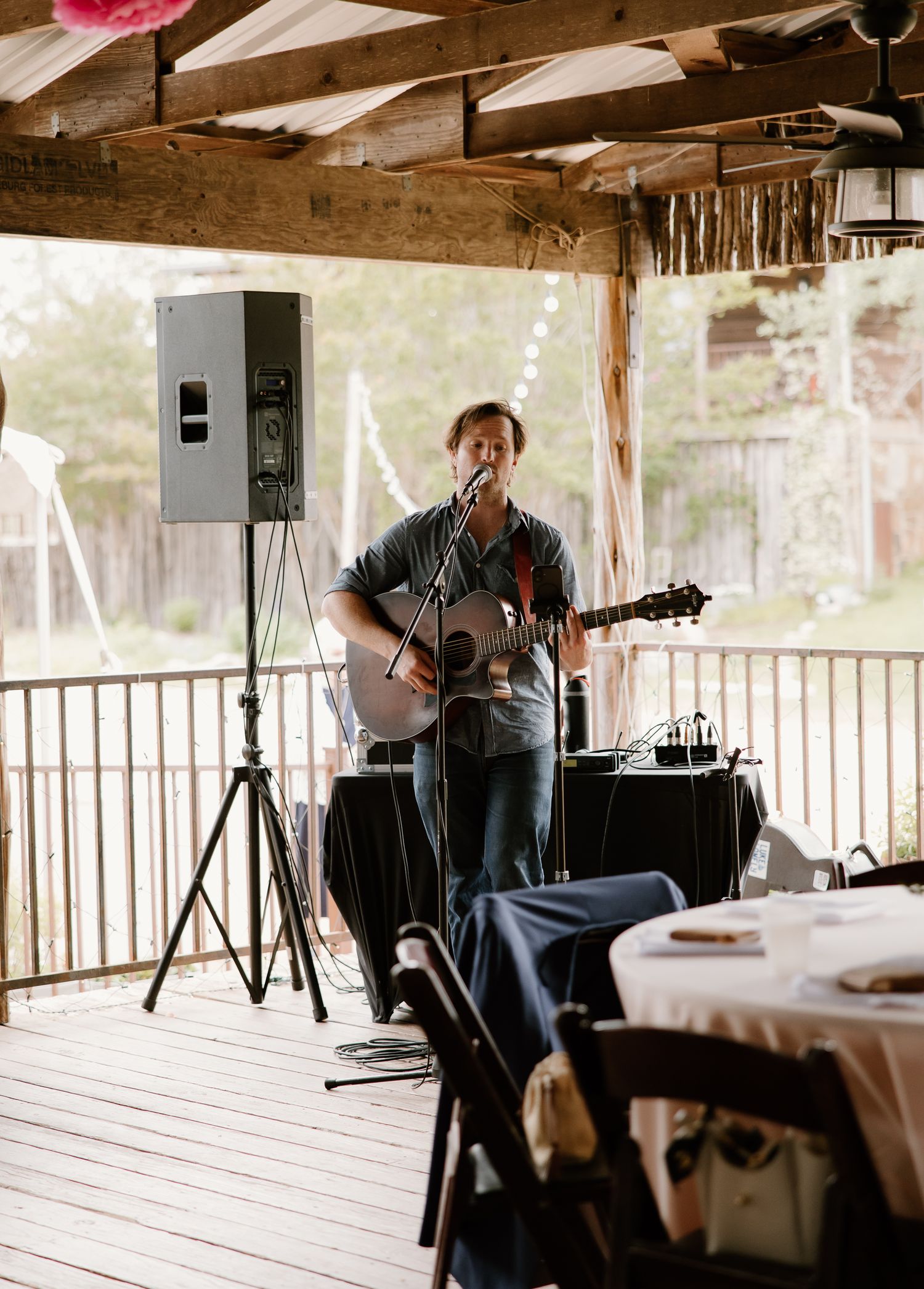 Acoustic guitarist performs under a wooden covered porch at a rustic outdoor wedding reception.