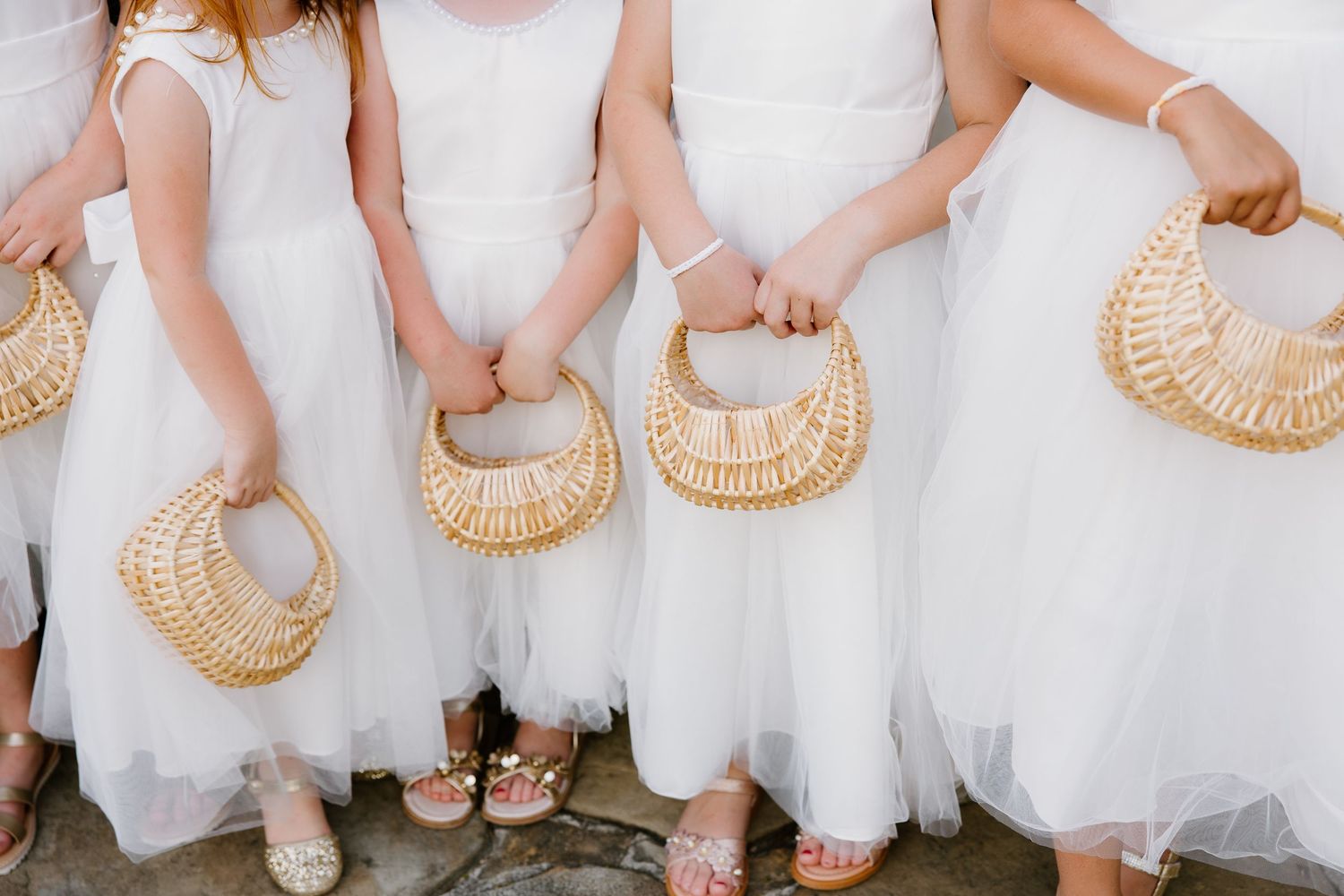 Four flower girls in white dresses holding woven rattan basket purses at a wedding ceremony.