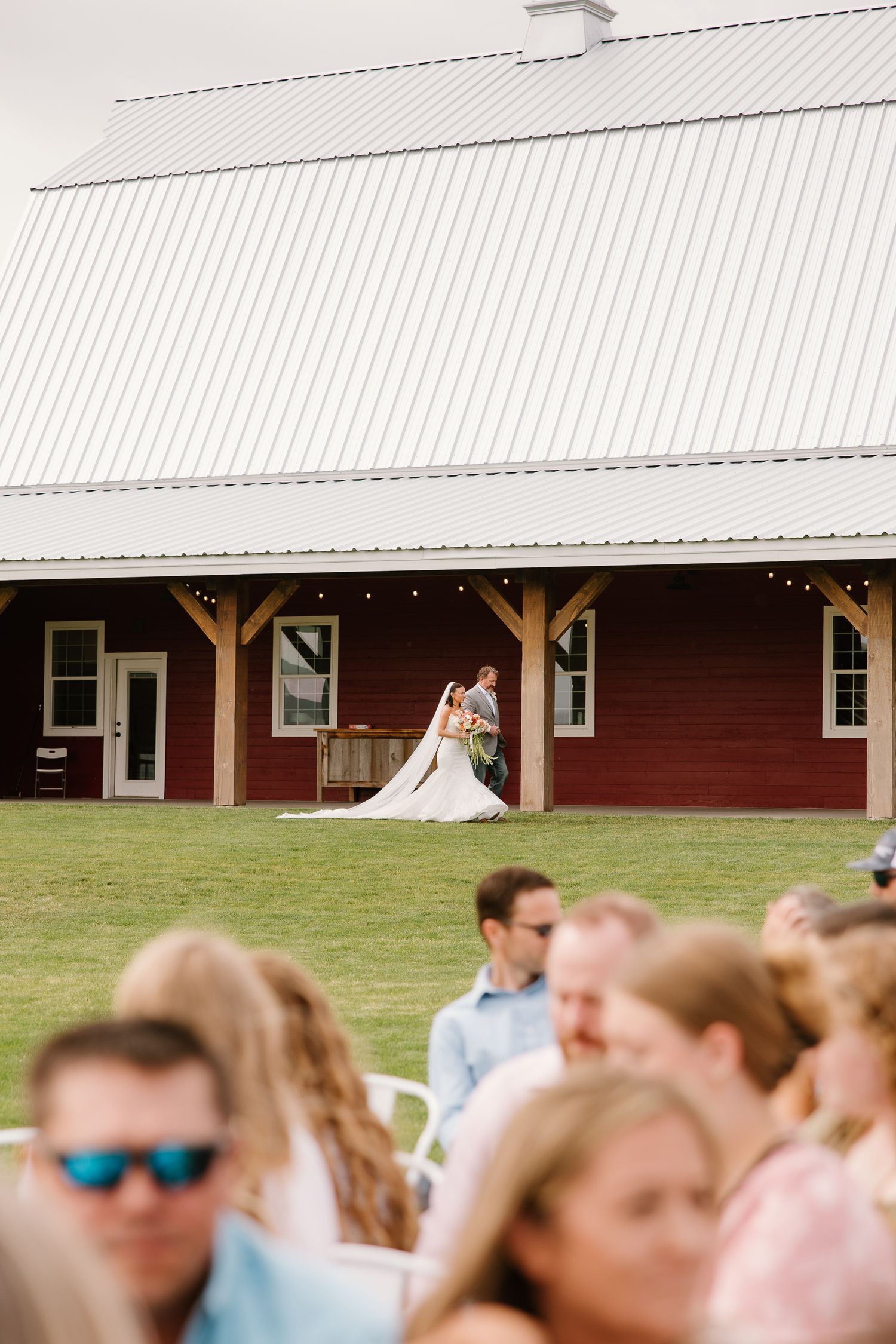 Bride and groom stand before a red barn venue while wedding guests sit in the foreground during an outdoor ceremony.