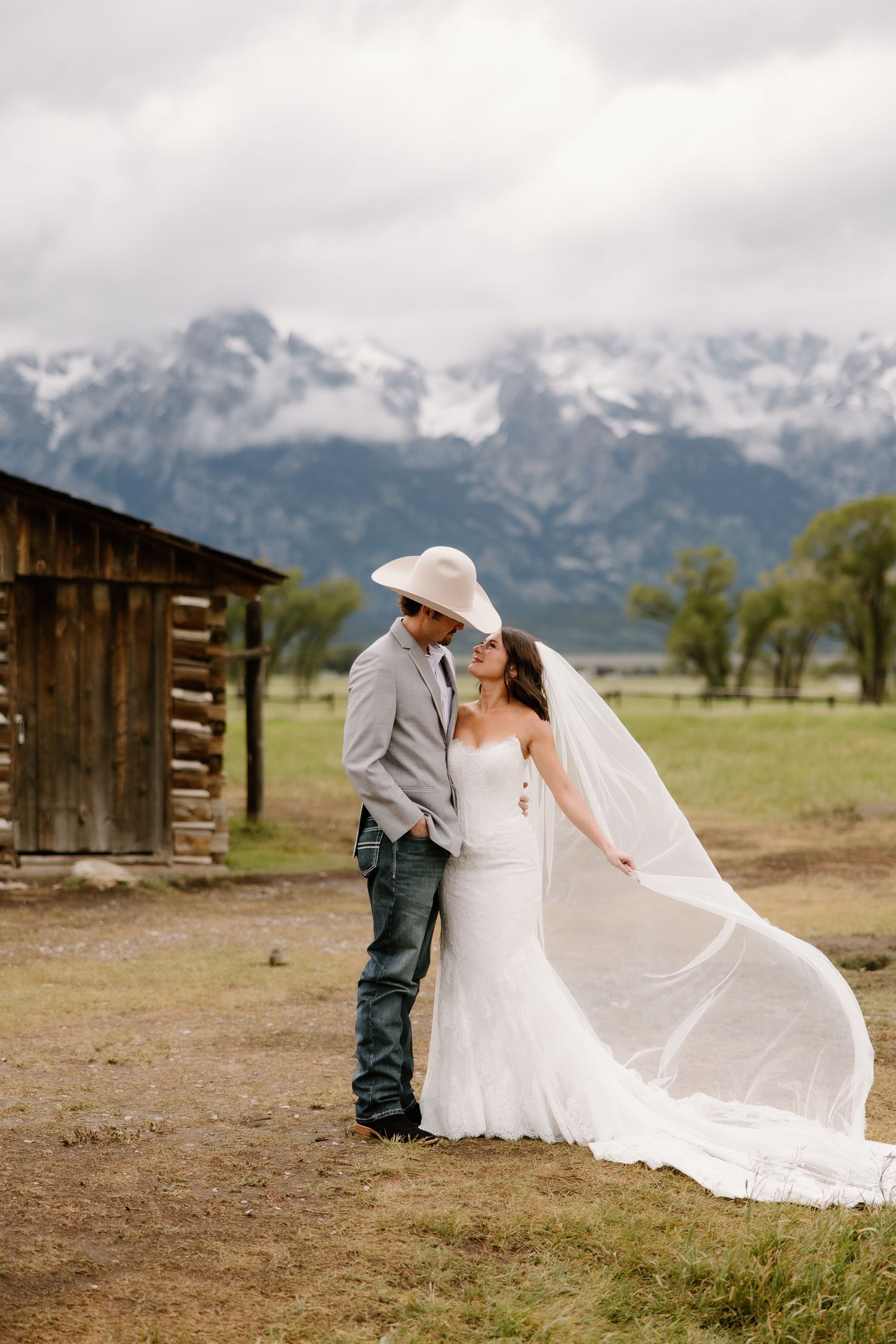 Bride and groom share a romantic kiss with snow-capped Grand Teton mountains and rustic log cabin backdrop.