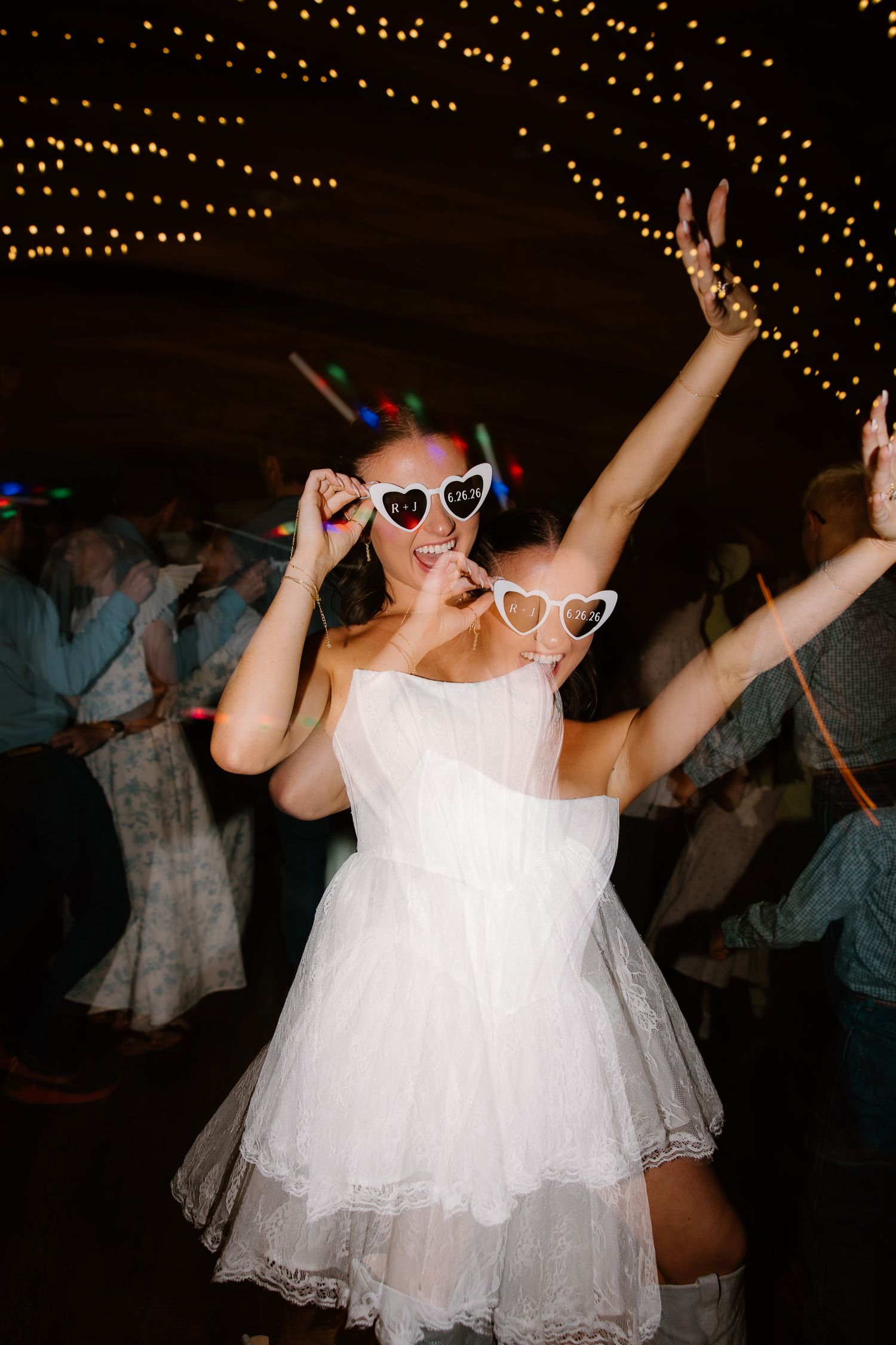 Bride in a short white dress dances joyfully wearing heart-shaped sunglasses under string lights at wedding reception.