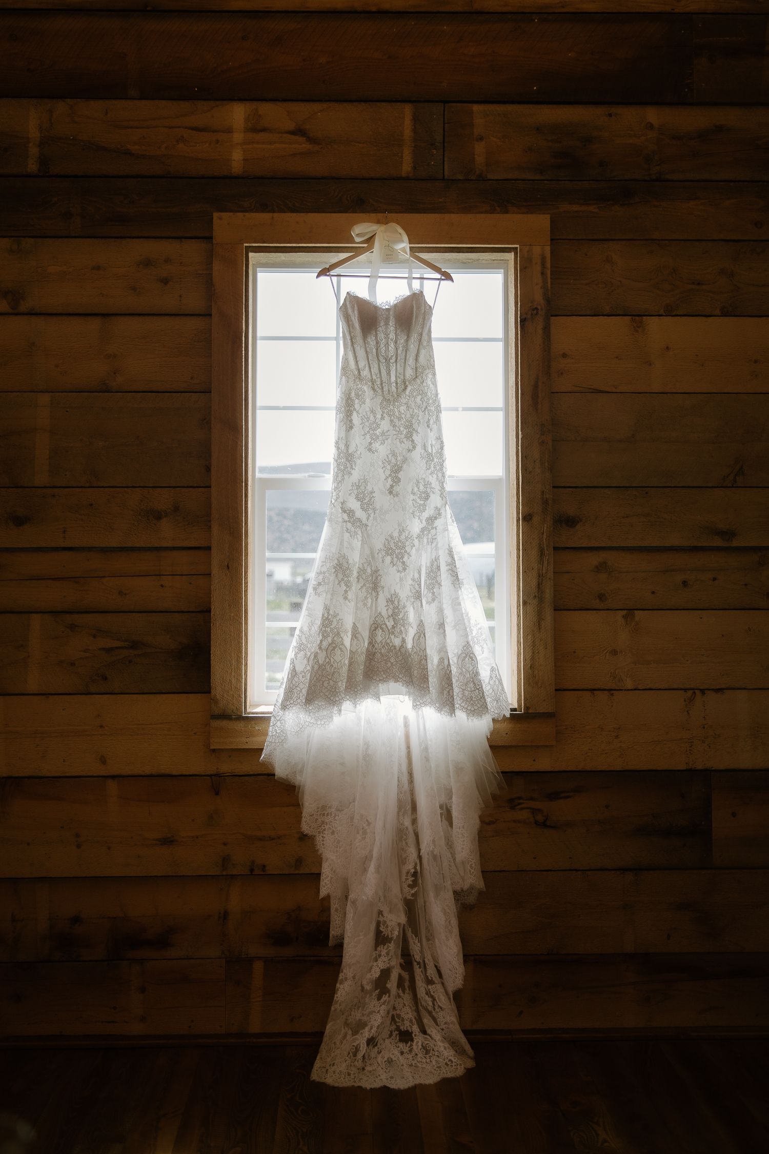 Elegant lace wedding dress hanging on a wooden hanger in front of a rustic cabin window with natural light.