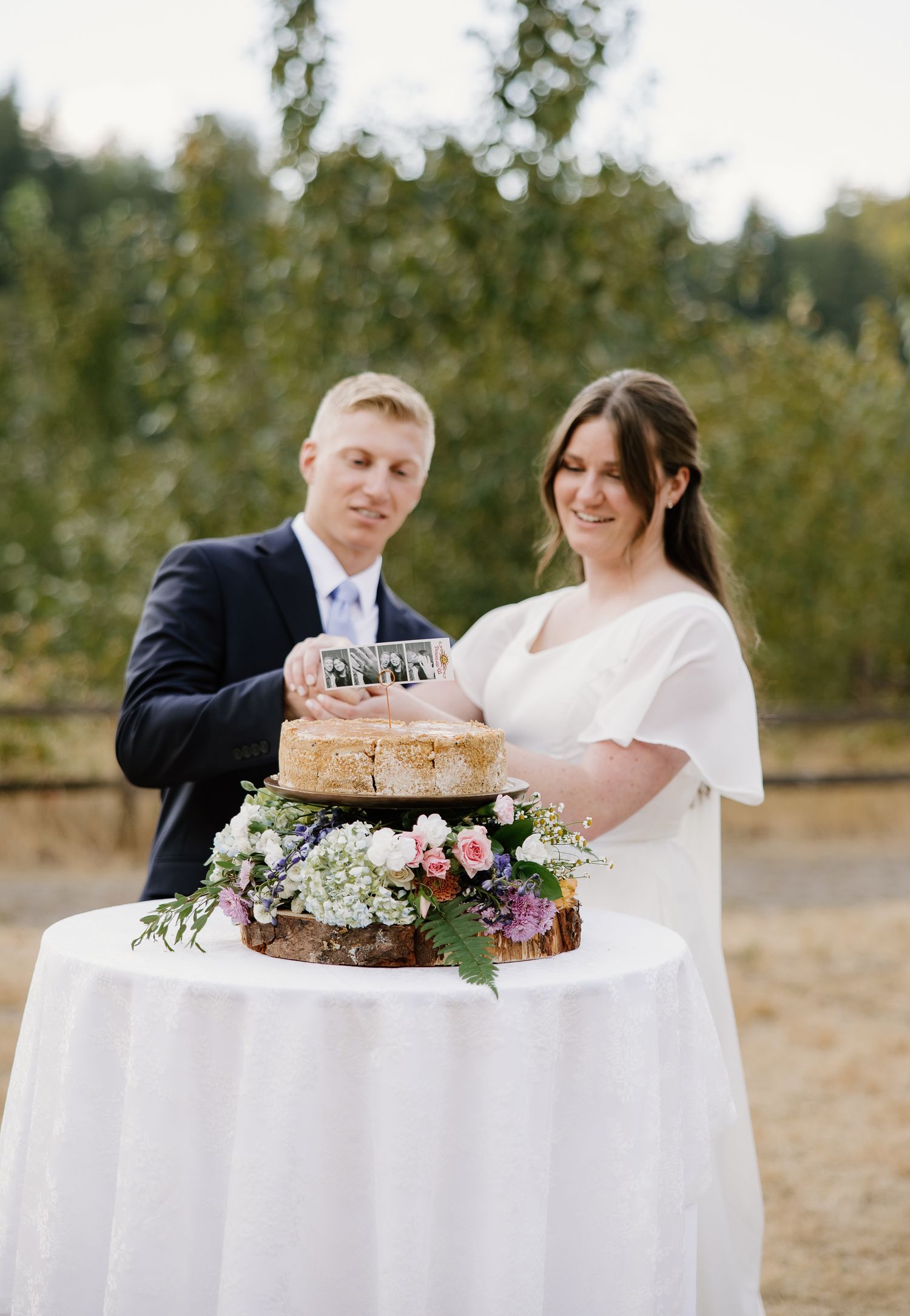Smiling bride and groom cut rustic naked wedding cake decorated with fresh flowers on white draped table.