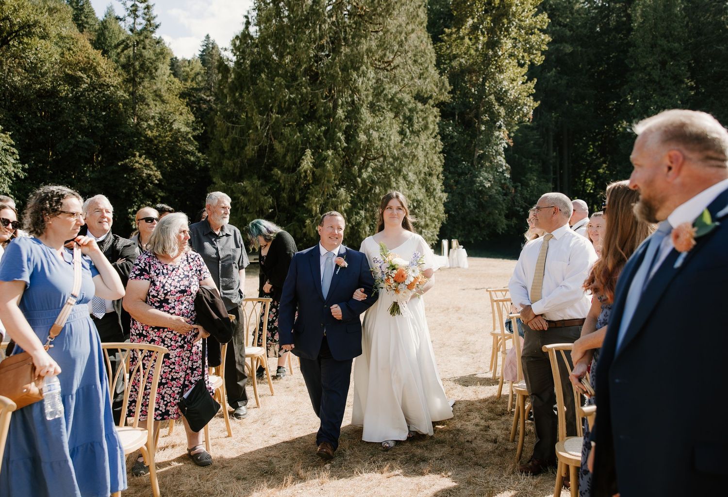 Bride walks down outdoor aisle with escort past seated wedding guests surrounded by tall trees.