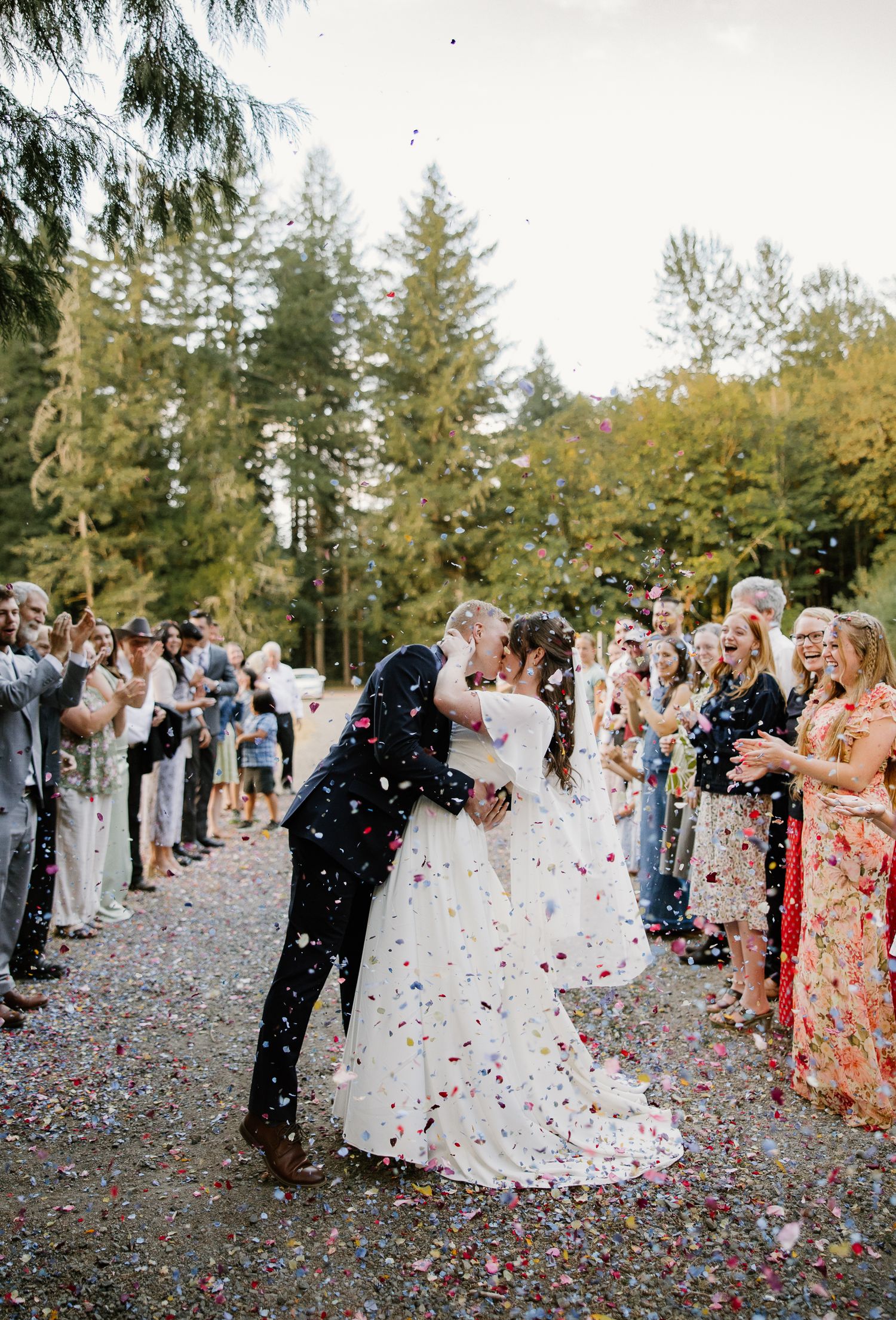 Bride and groom kiss during colorful confetti send-off surrounded by cheering wedding guests outdoors.