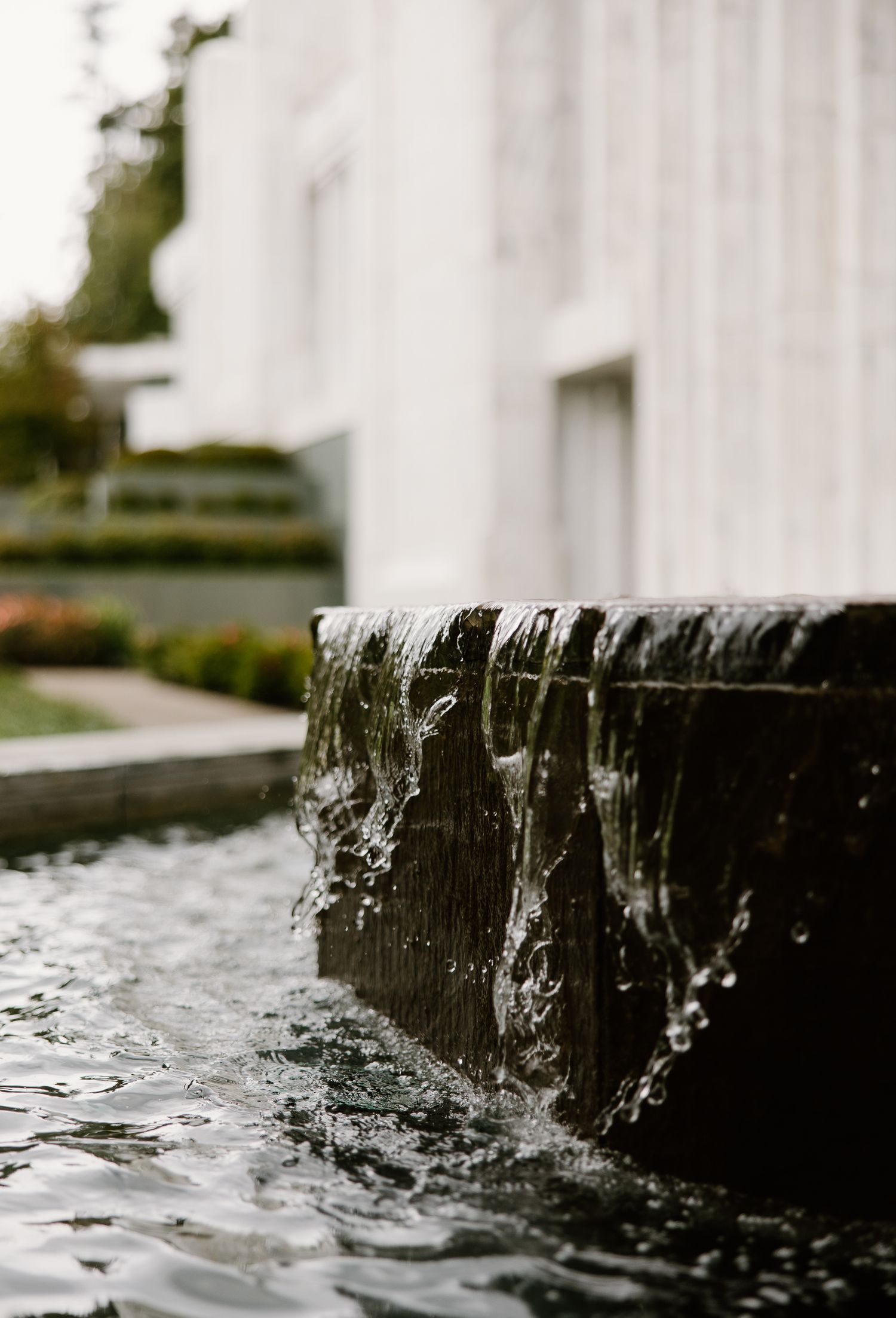 Water cascading over a dark stone fountain into a serene pool, with a white modern building in the soft background.