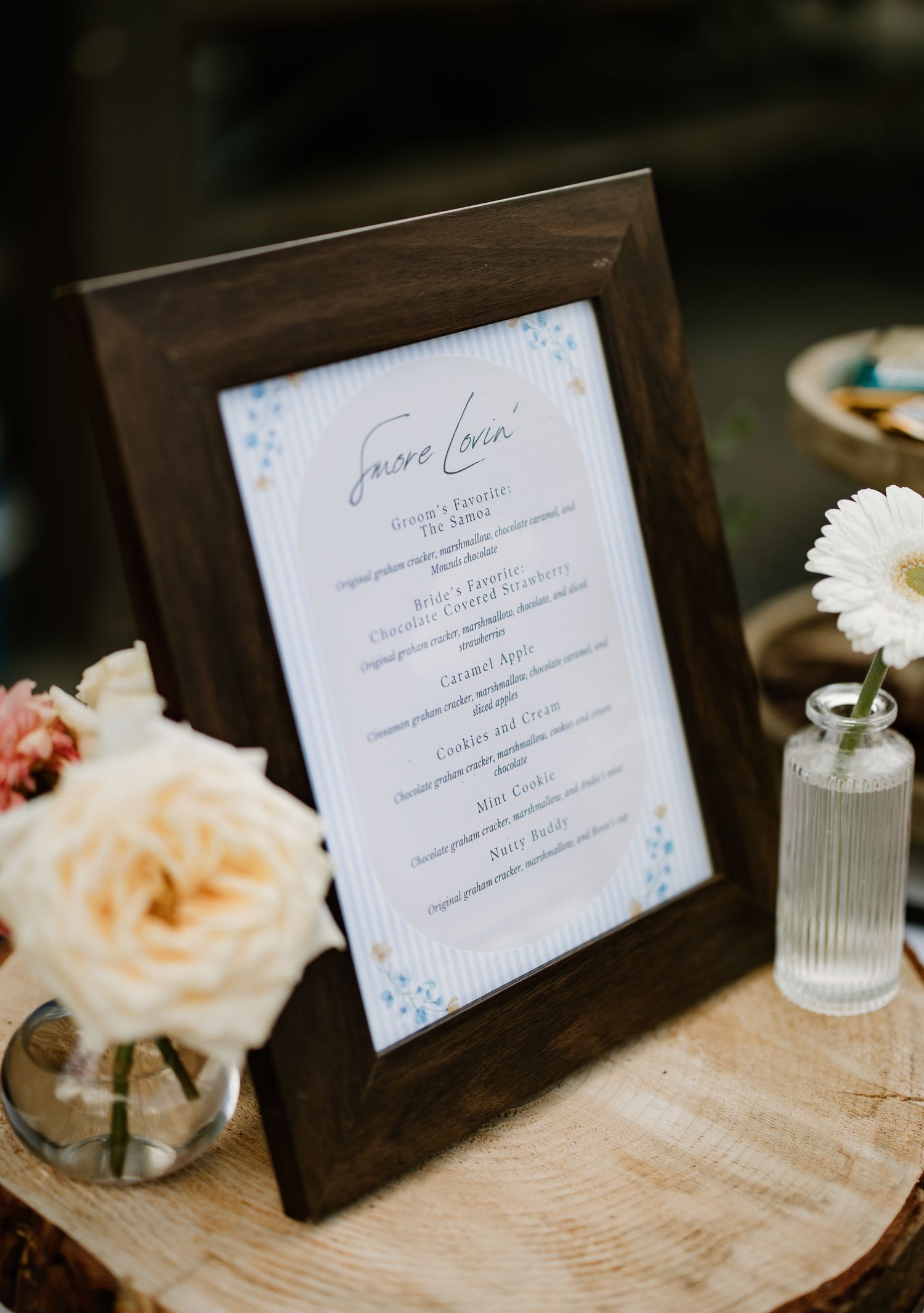 Framed wedding menu displayed on wood slice with white rose and daisy floral arrangements nearby.