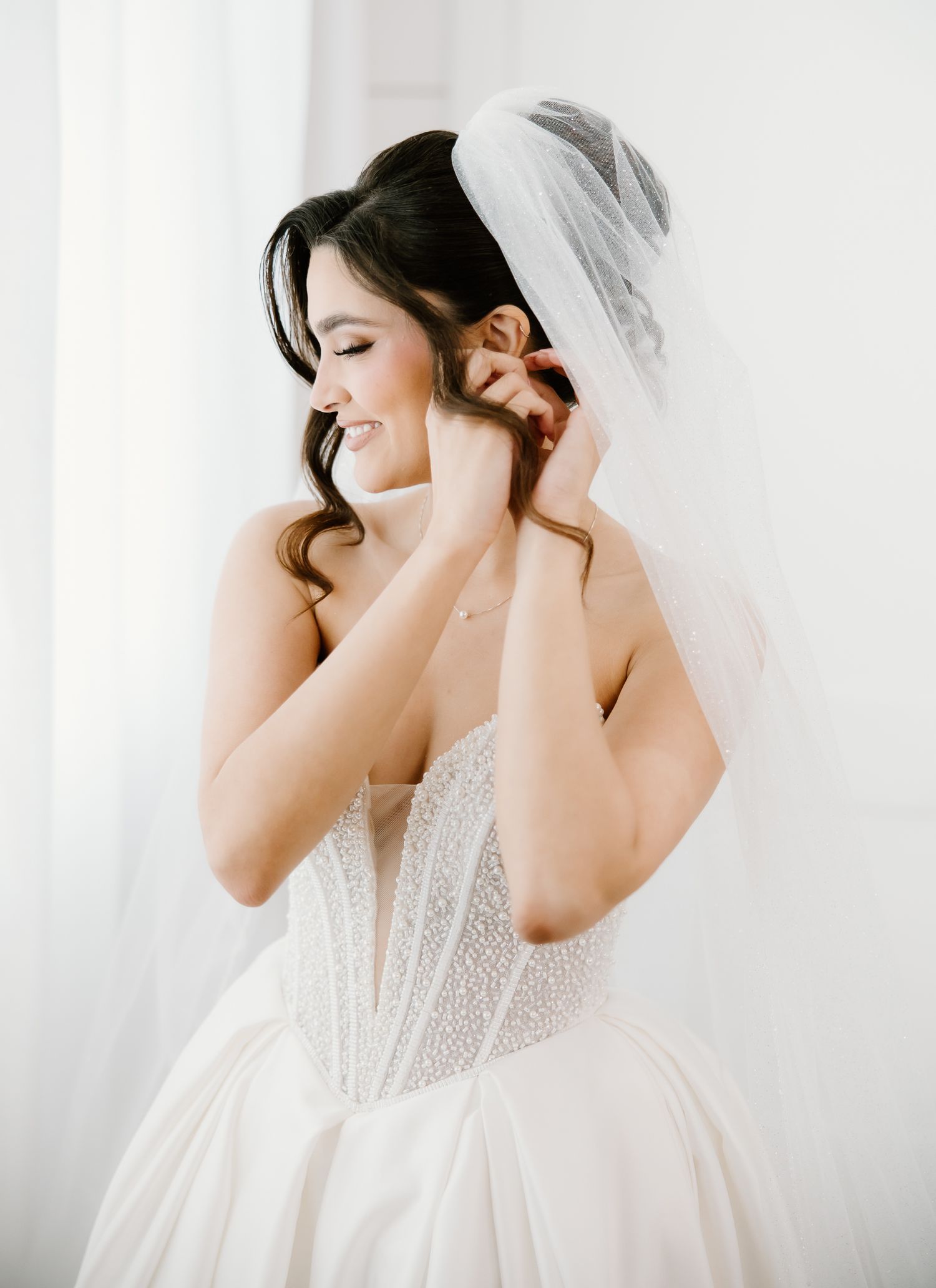 Radiant bride in beaded corset ballgown adjusts earring while wearing a flowing veil.