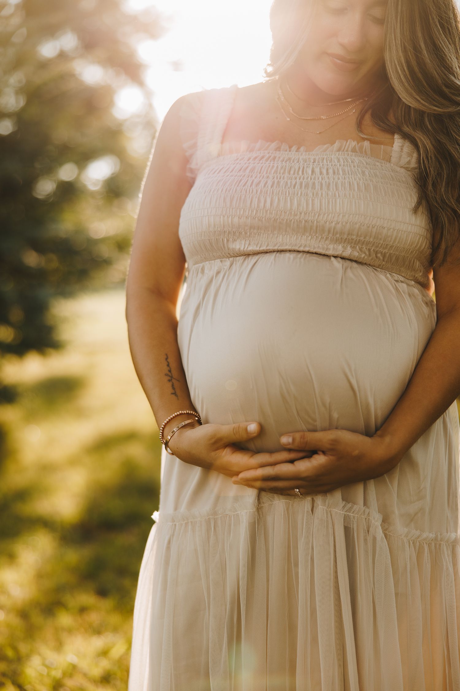 Pregnant woman in cream smocked dress cradles baby bump in warm golden sunlight outdoors.