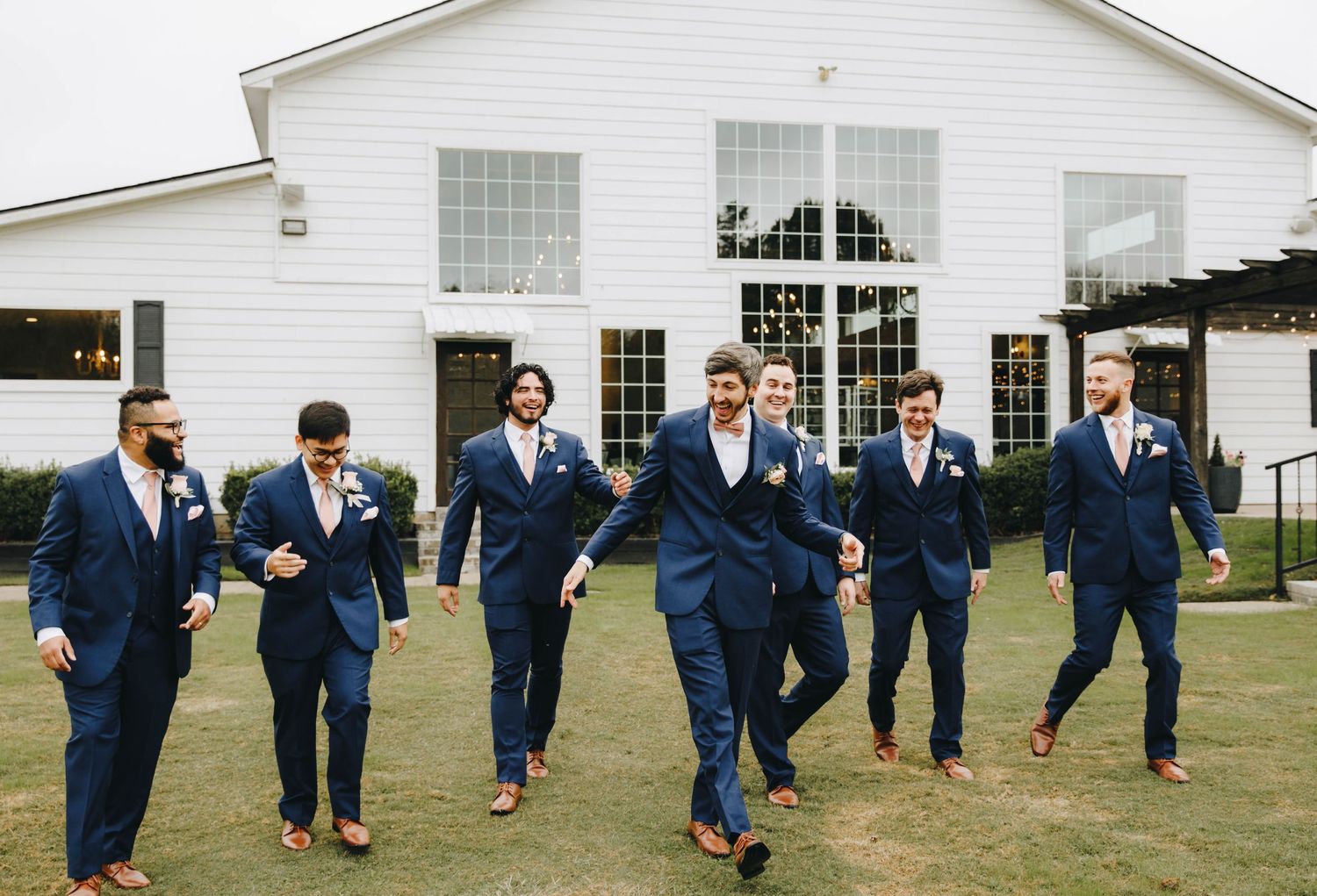 Groomsmen in navy blue suits walking and laughing together outside a white wedding venue.