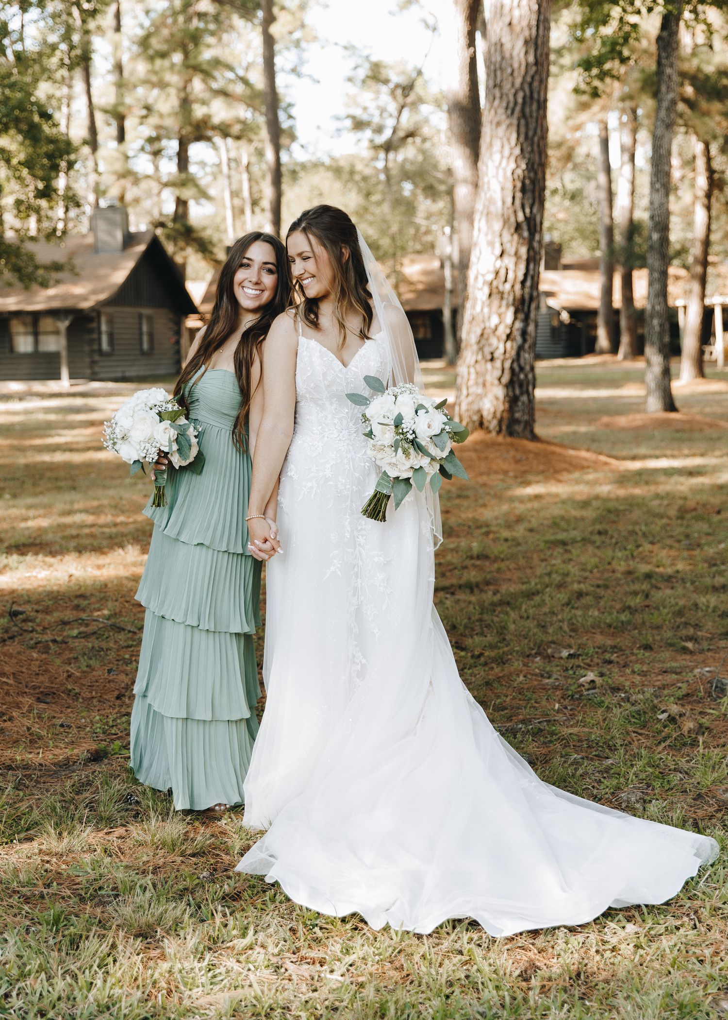 Bride in white gown and bridesmaid in sage green tiered dress holding bouquets outdoors among pine trees.