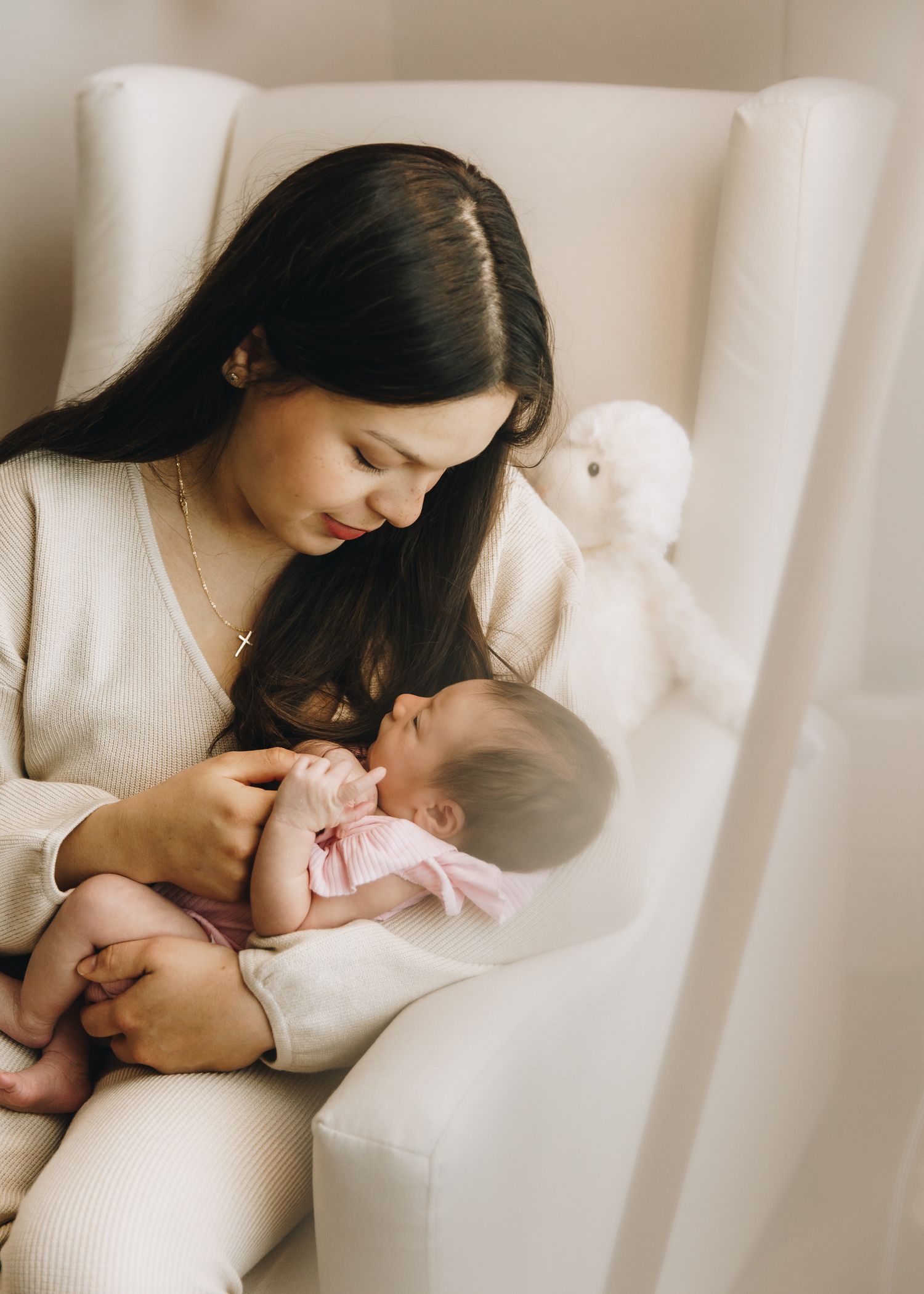 Dark-haired mother in cream outfit gazing lovingly at newborn baby in white nursery chair.