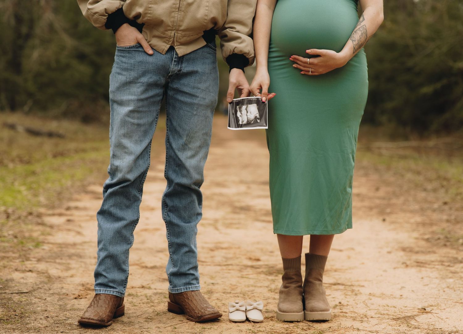 Couple holding ultrasound photo and baby shoes on dirt path during outdoor maternity announcement session.