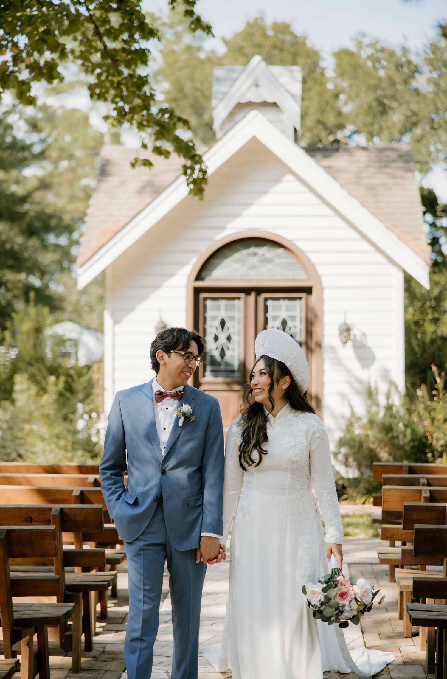 Couple in wedding attire hold hands and smile outside a charming white chapel surrounded by trees.