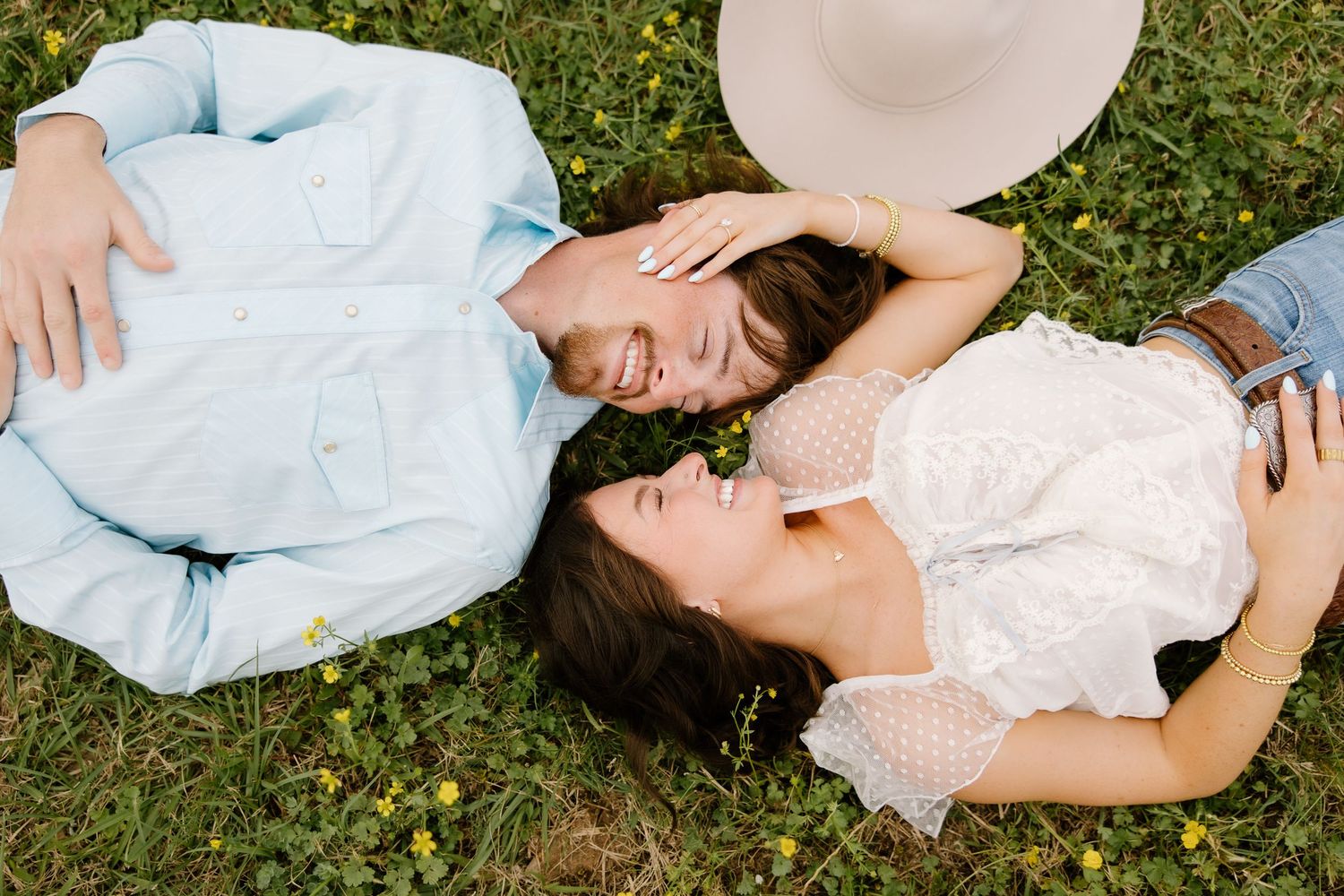 Couple laughing while lying on green grass with wildflowers during engagement session.