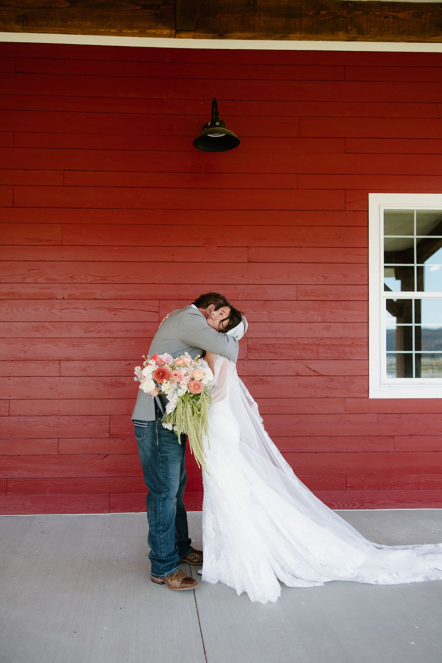 Bride and groom share a tender embrace against a red barn wall, bridal gown train flowing gracefully.