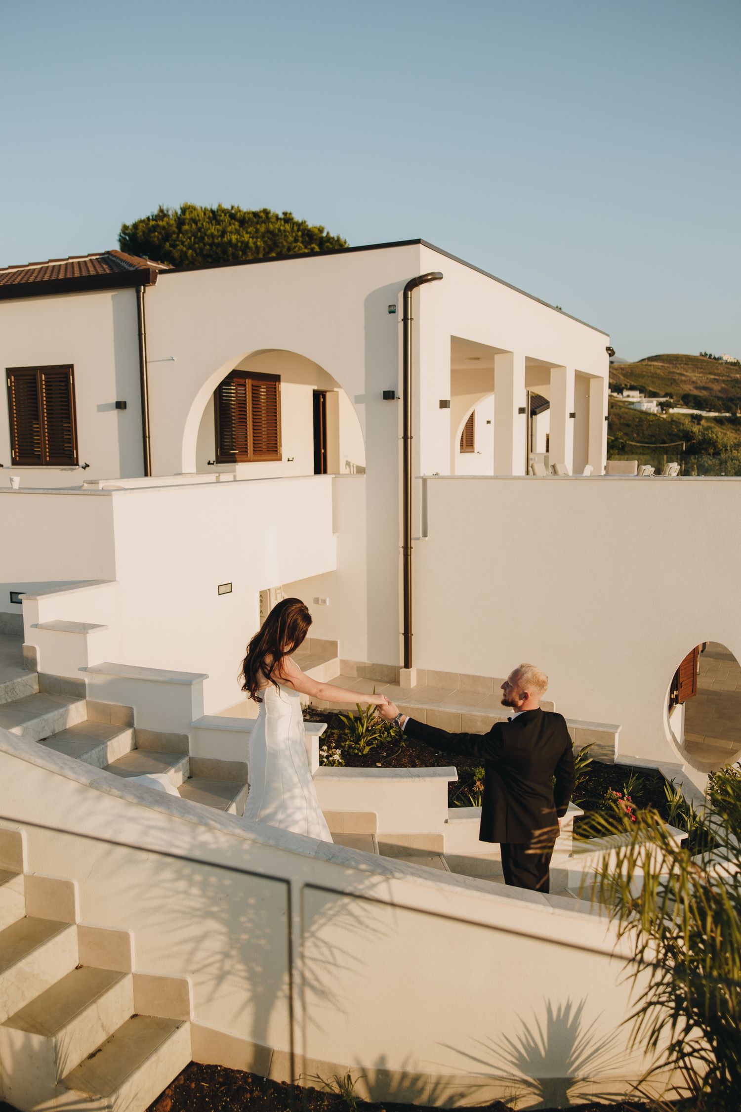 Bride and groom hold hands on white Mediterranean villa steps in golden hour light.