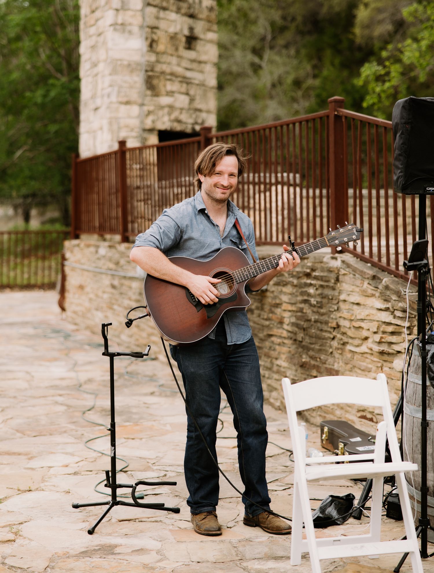 Smiling musician playing acoustic guitar outdoors at a wedding venue with stone fireplace backdrop.
