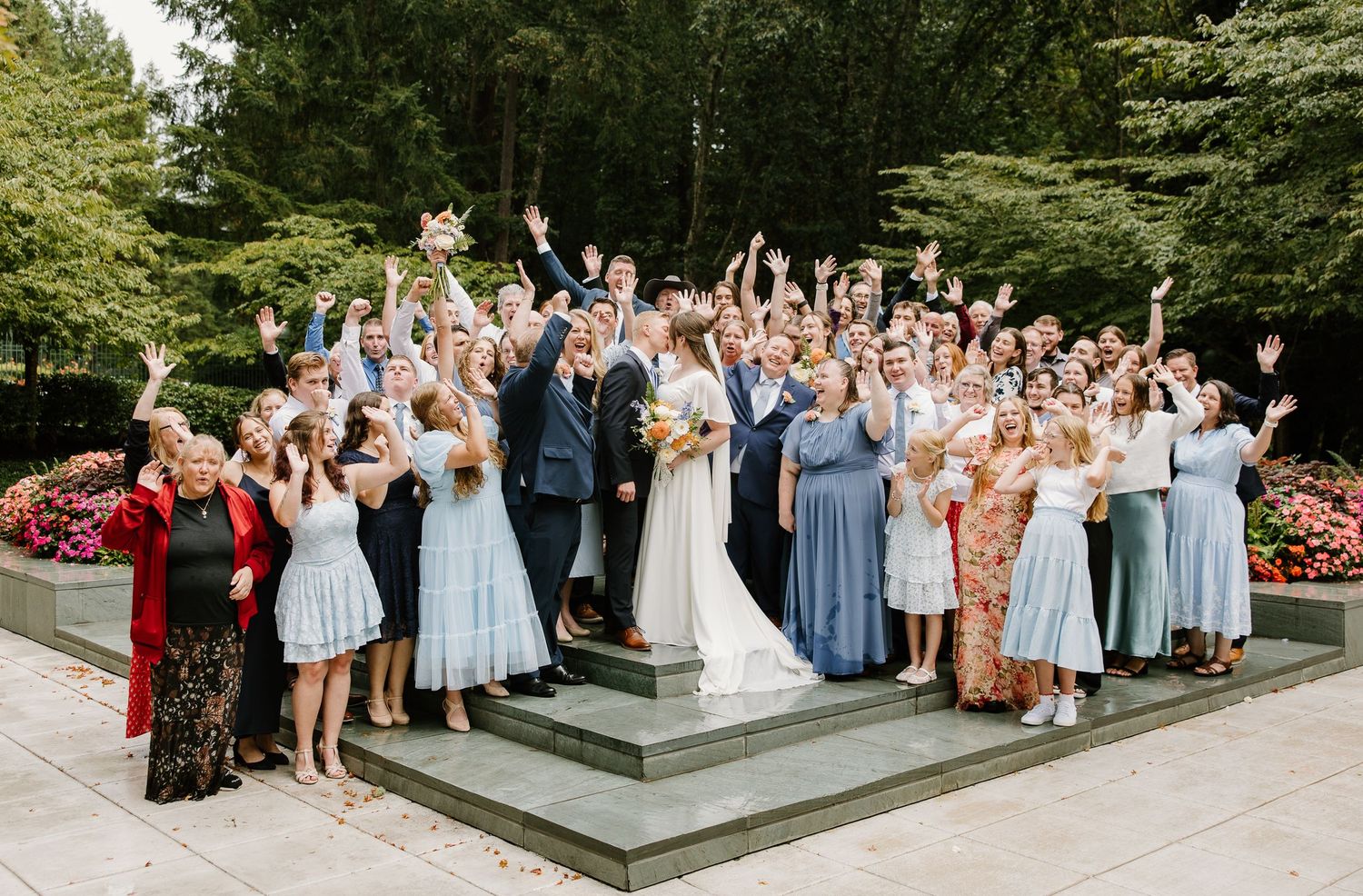 Large wedding party celebrating outdoors on steps, bride and groom surrounded by guests with hands raised, lush green trees behind.