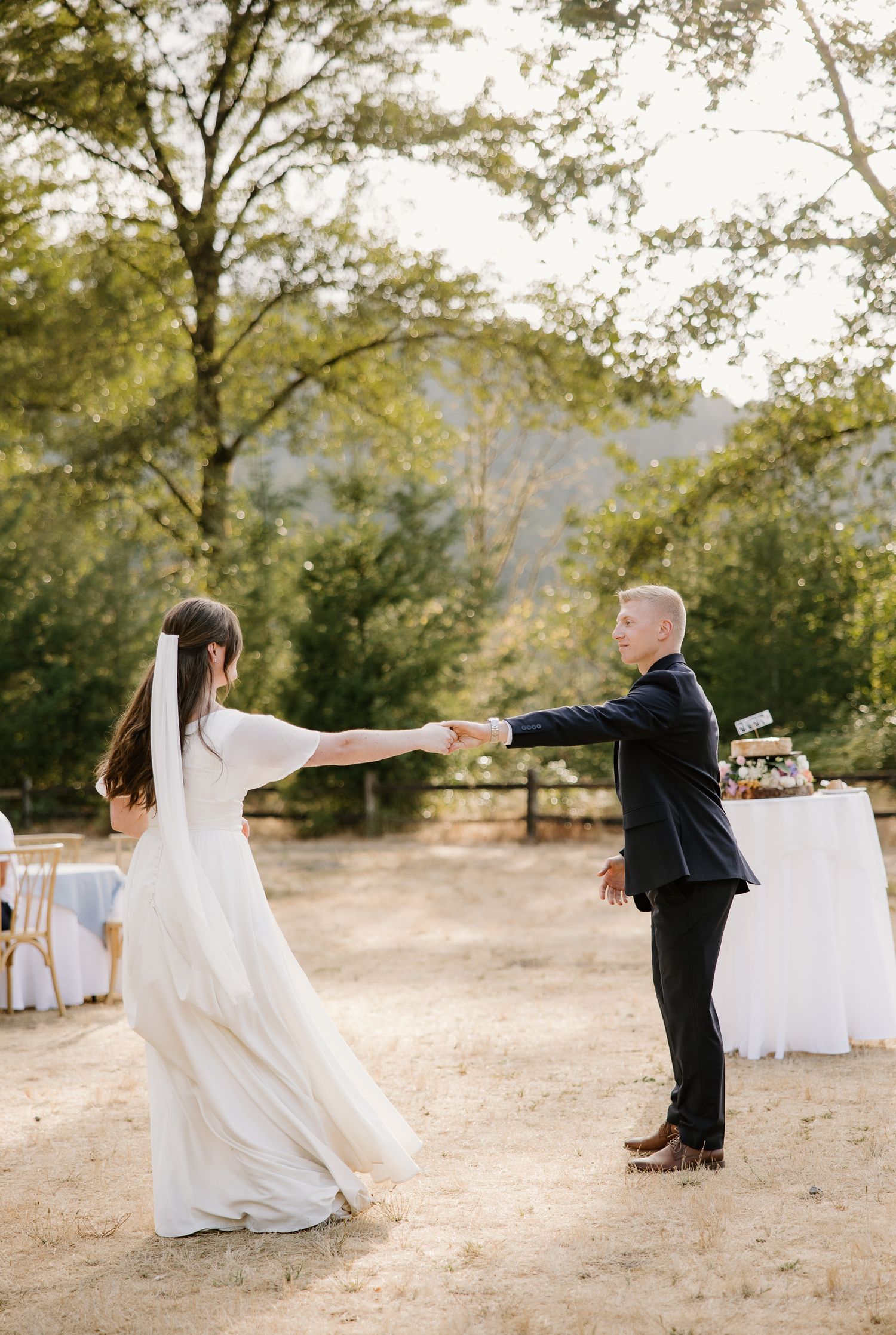 Bride and groom share first dance outdoors on sandy ground surrounded by lush green trees at a wedding reception.
