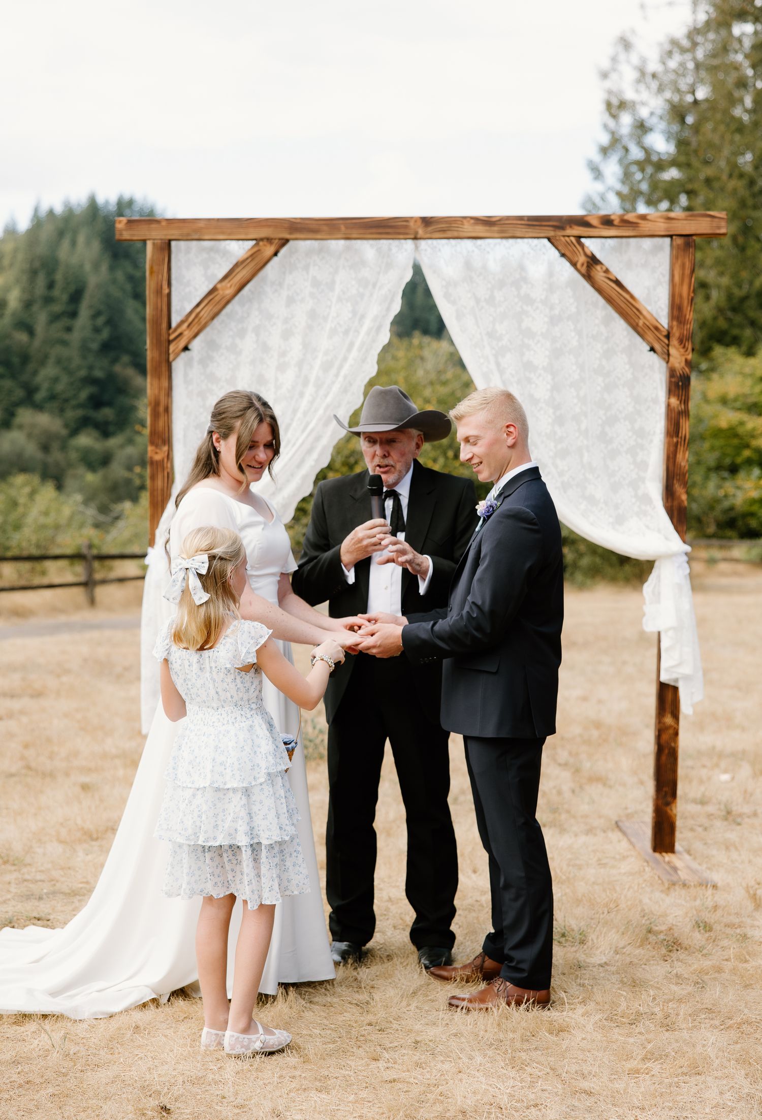 Bride, groom, and flower girl participate in a unity ceremony under a rustic wooden arch.