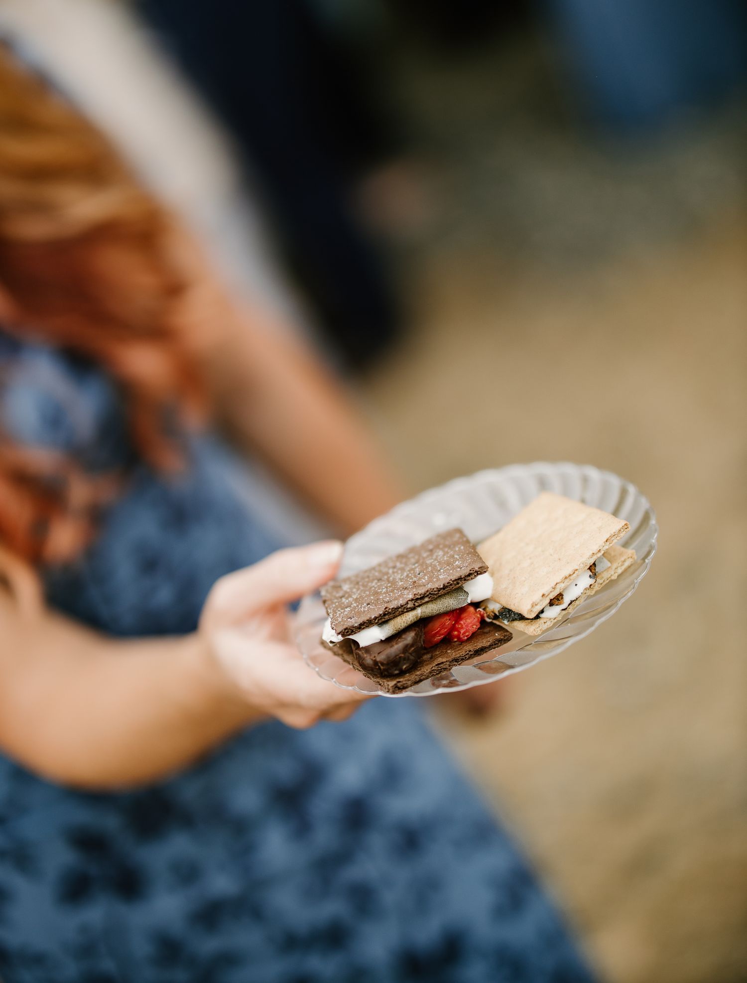 Woman in blue floral dress holds a plate with a strawberry s'more at a wedding reception.