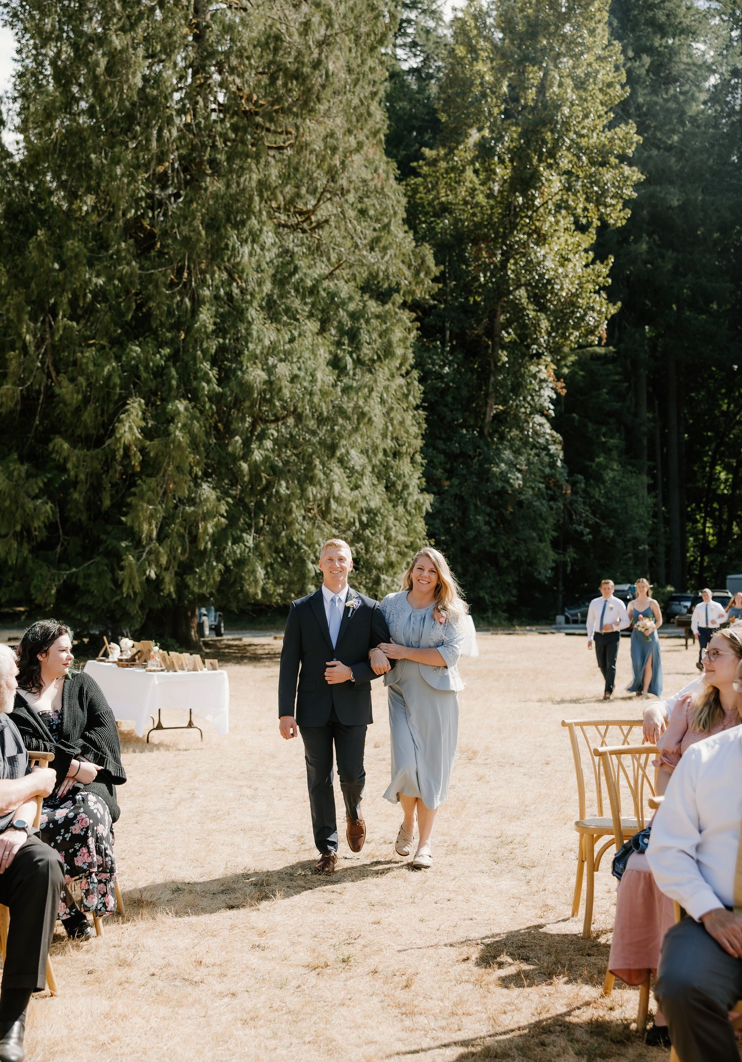 A couple walks down the outdoor wedding aisle surrounded by tall evergreen trees and seated guests.