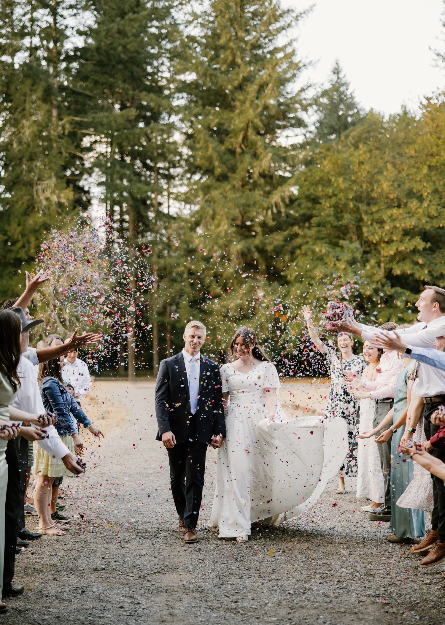 Newlyweds walk through colorful confetti shower as smiling wedding guests celebrate their exit outdoors.