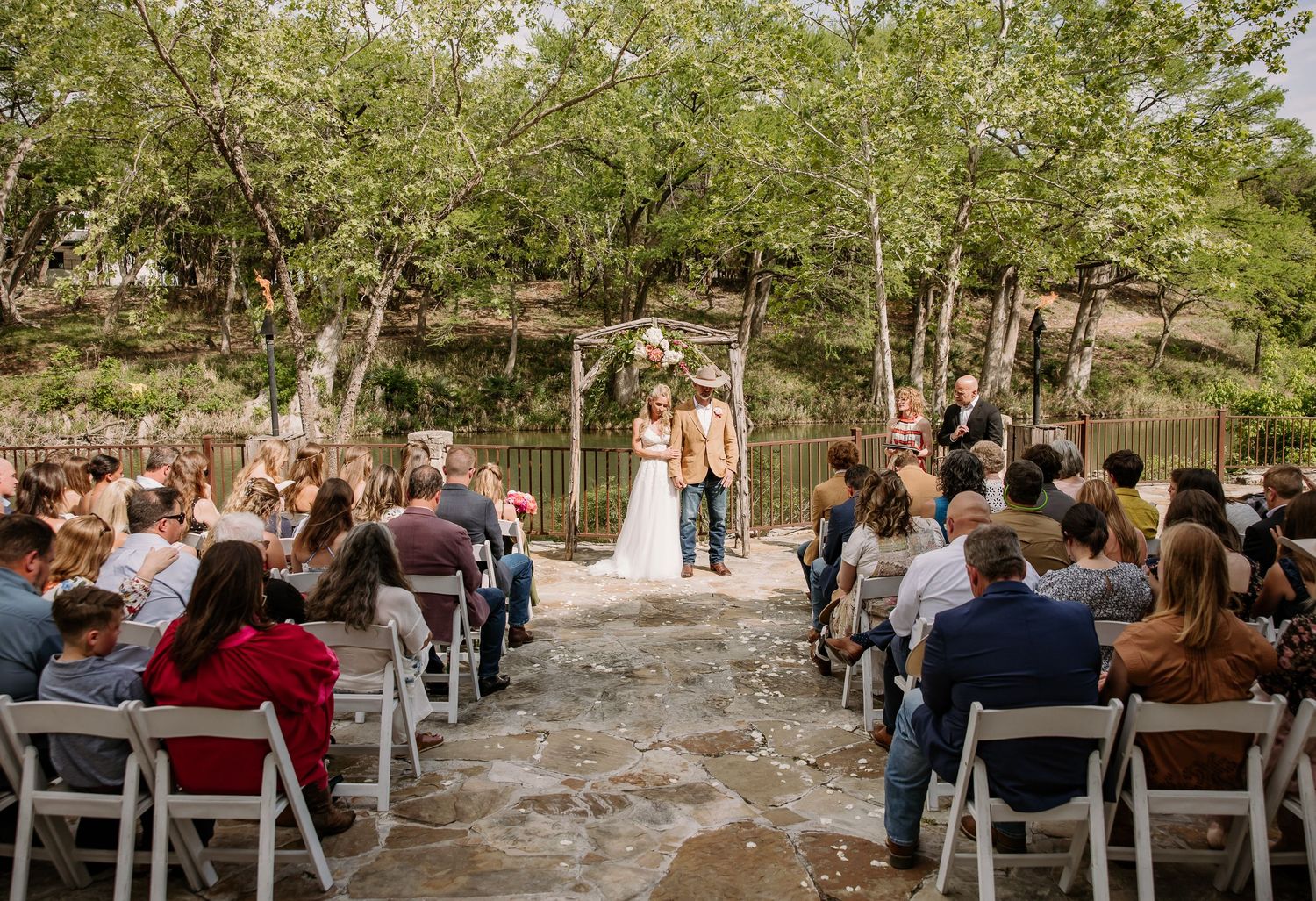 Outdoor rustic wedding ceremony with guests seated in white chairs as couple exchanges vows under floral arch.