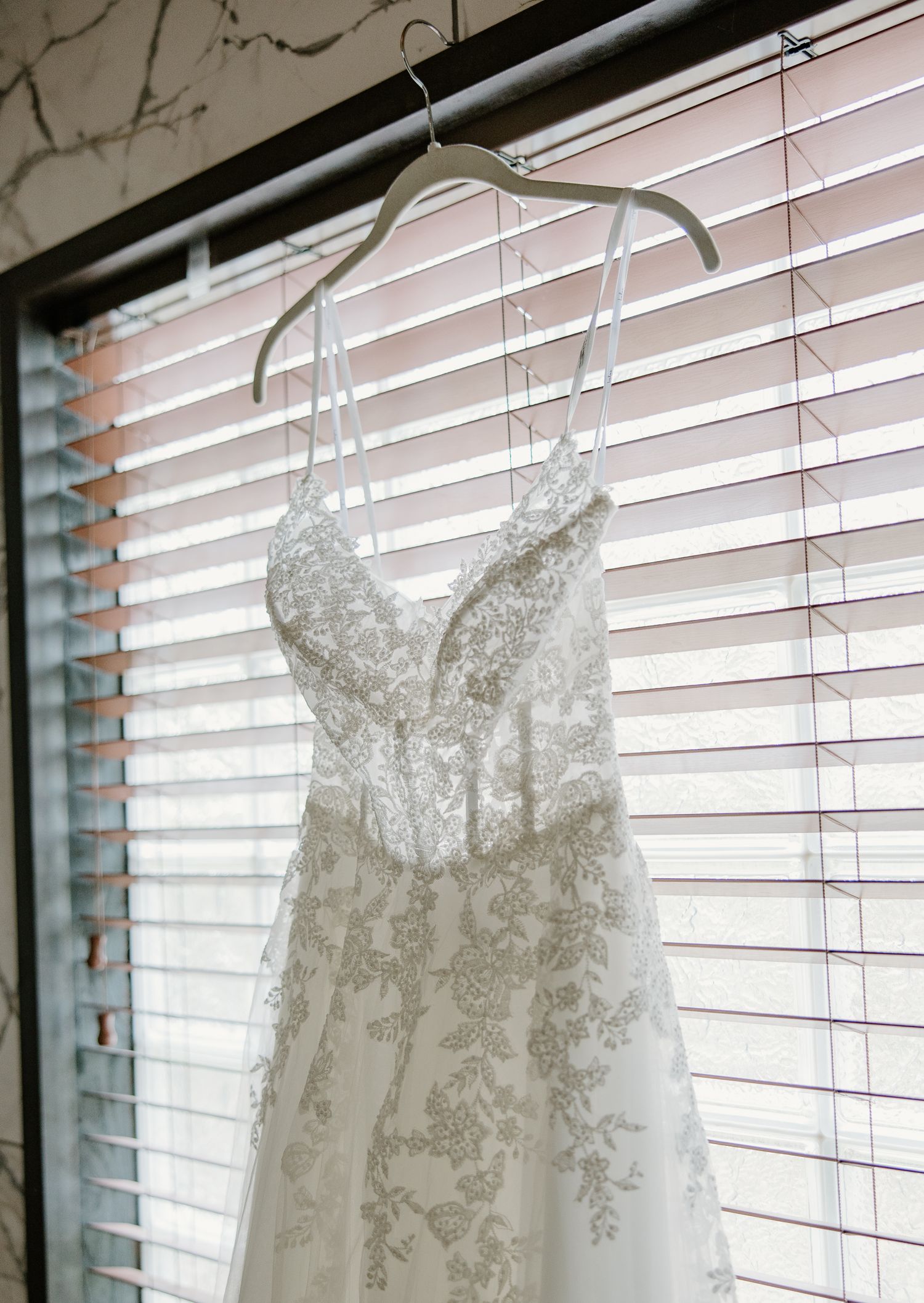 A delicate lace wedding dress with spaghetti straps hangs on a hanger in front of window blinds.
