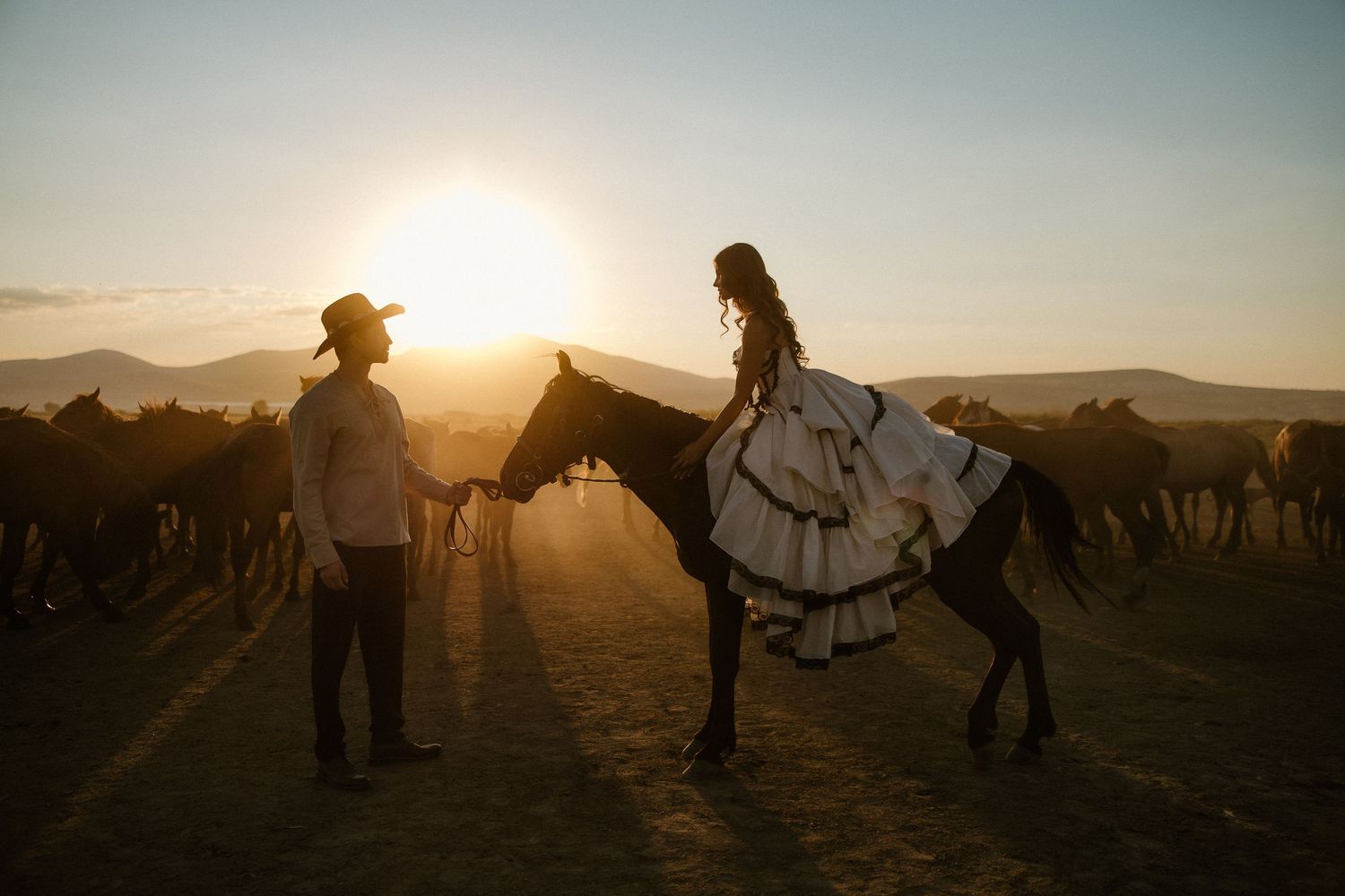 Couple in traditional attire at sunset, woman in white ruffled dress on horseback, man in cowboy hat, golden light.